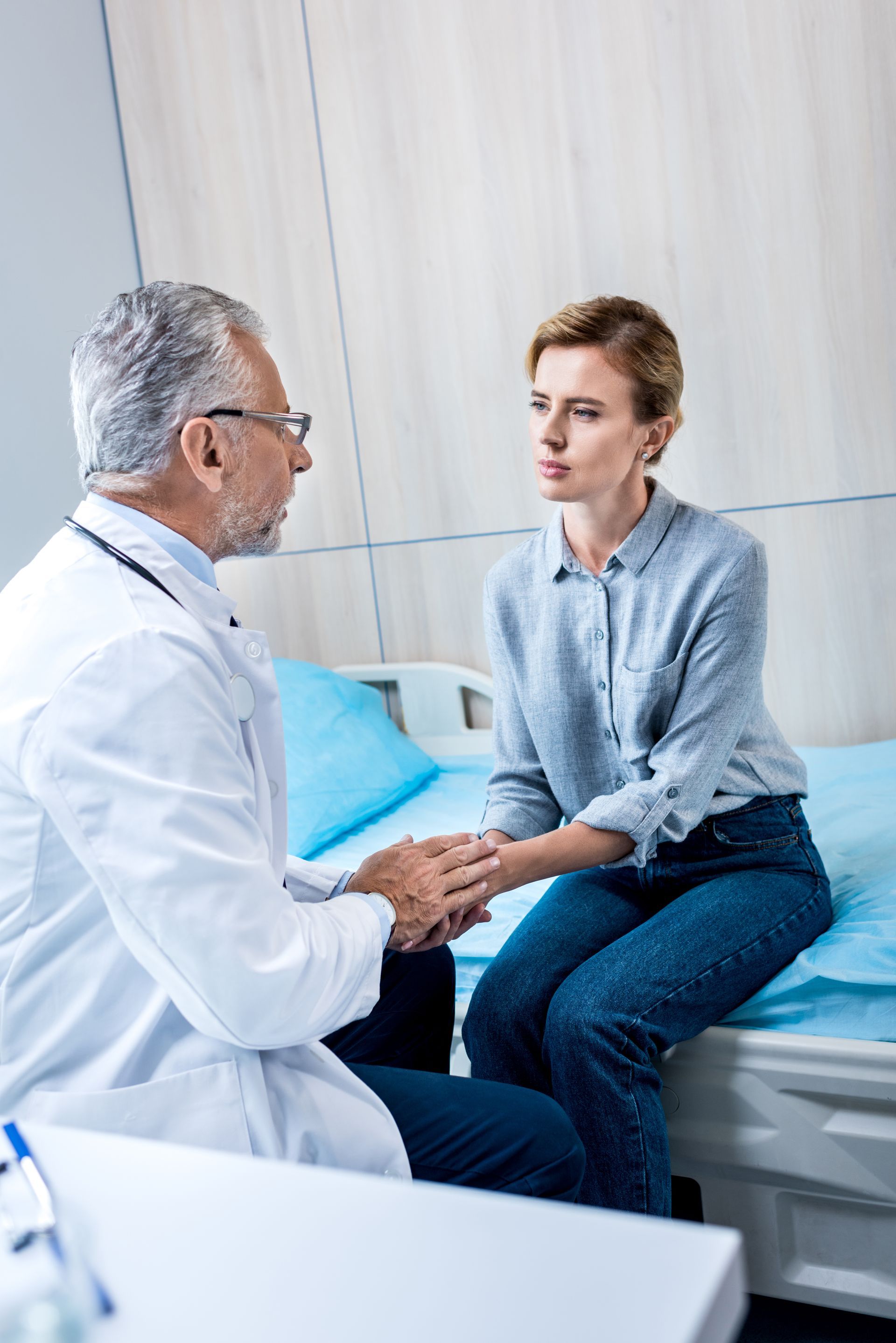 Doctor speaking with patient in a clinic room, seated beside a bed, holding hands
