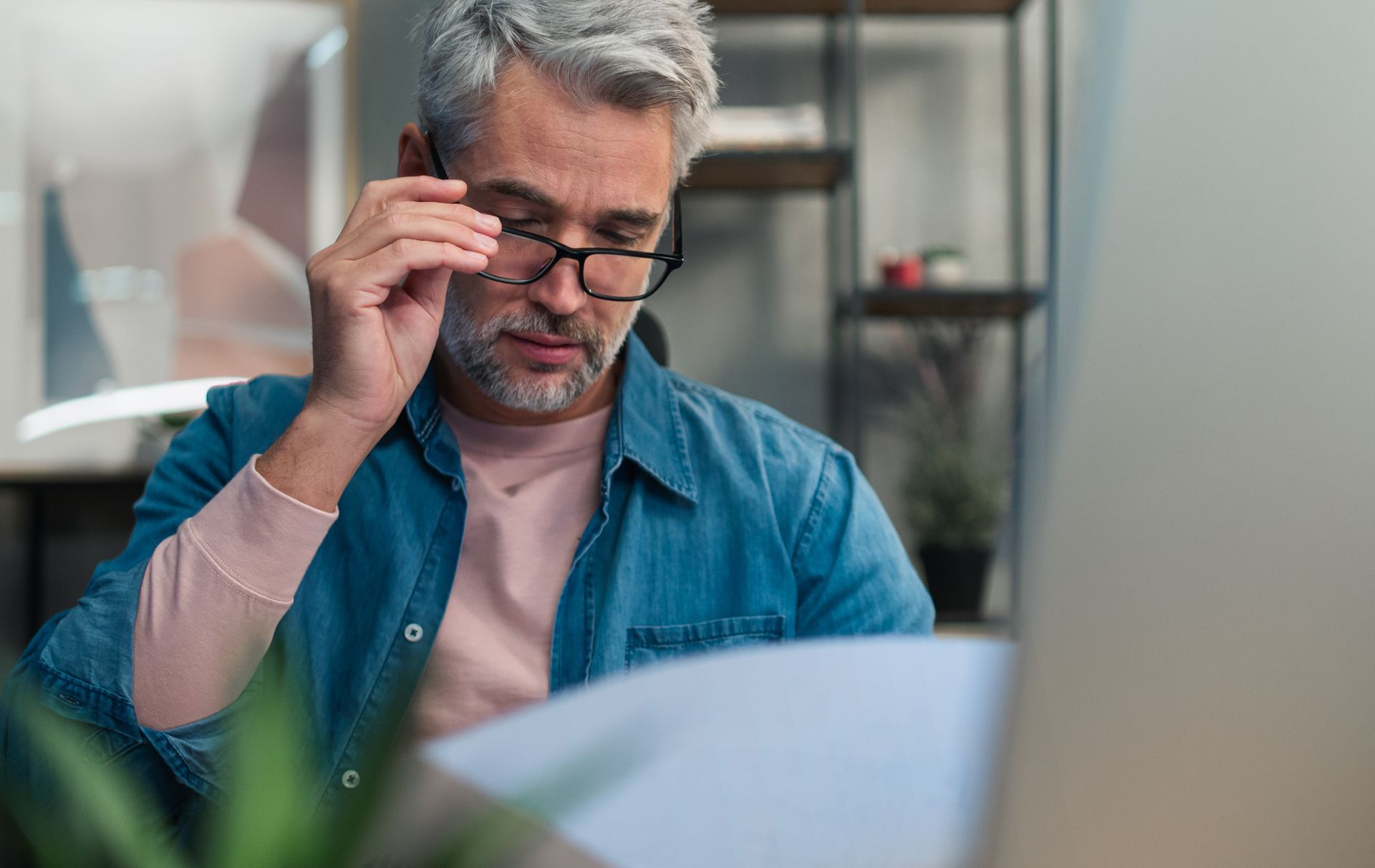 Man adjusting glasses while reading papers at a desk, with a blurred indoor background.