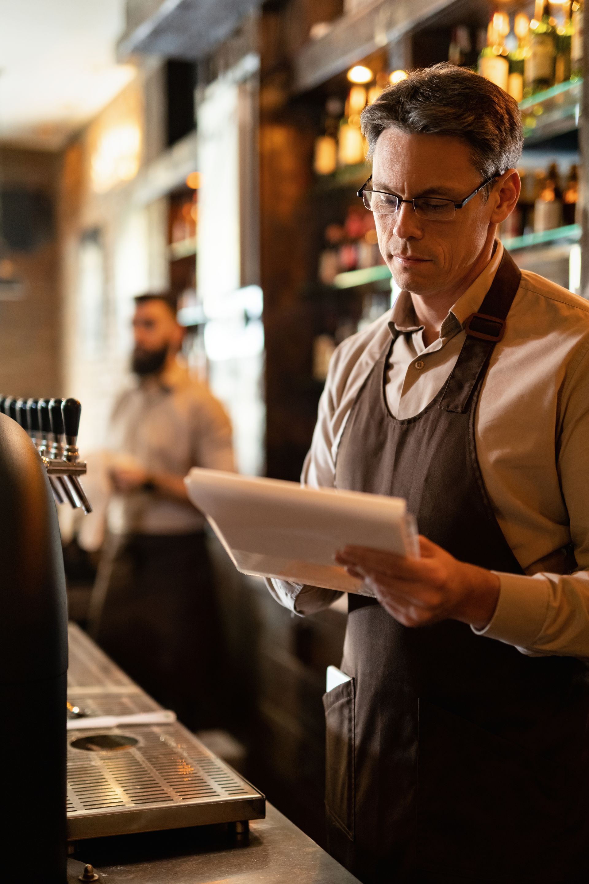 Bartender in a dark apron reading a tablet behind a bar, with bottles and warm lighting in the background
