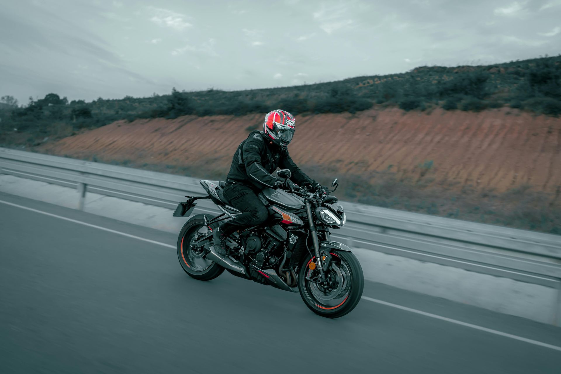 Motorcyclist in black gear riding a sport bike on a road beside a hillside under cloudy skies