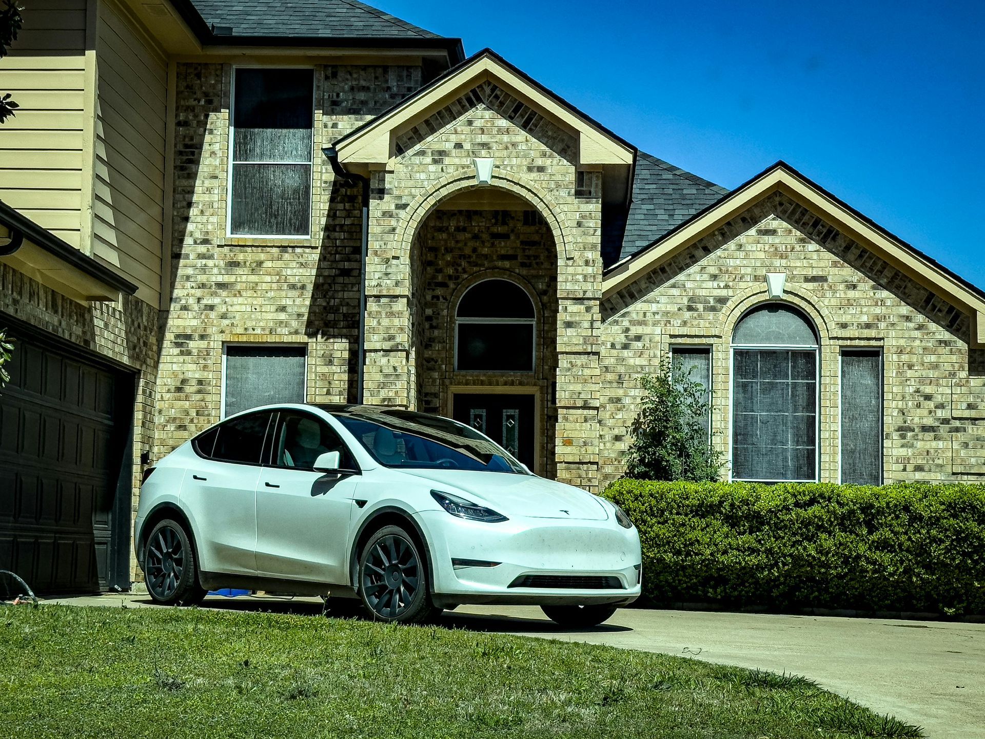 White electric car parked in front of a brick house with arched entry and trimmed hedges