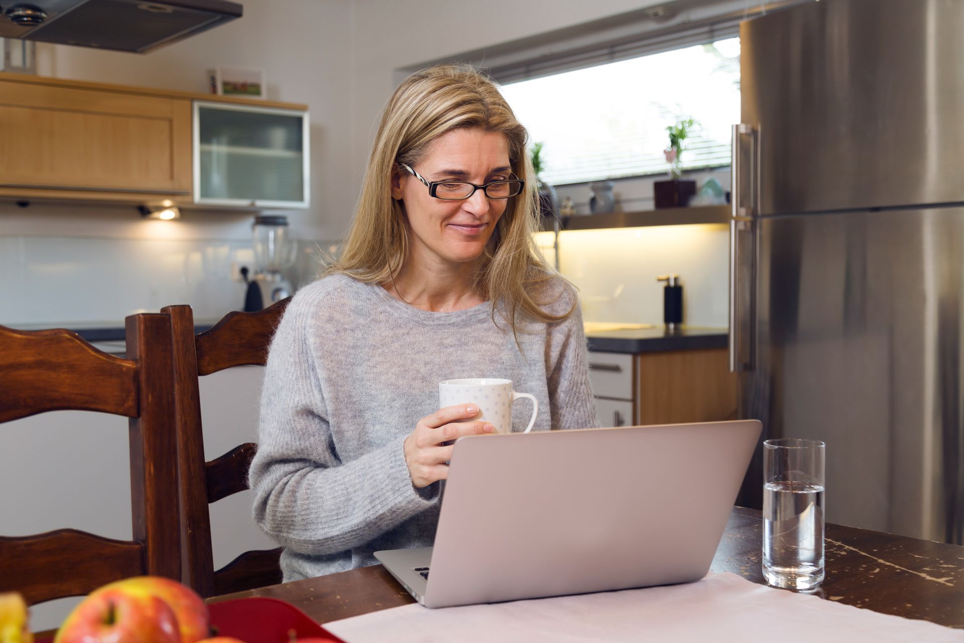 Woman using a laptop at a kitchen table, holding a mug in a bright home kitchen.