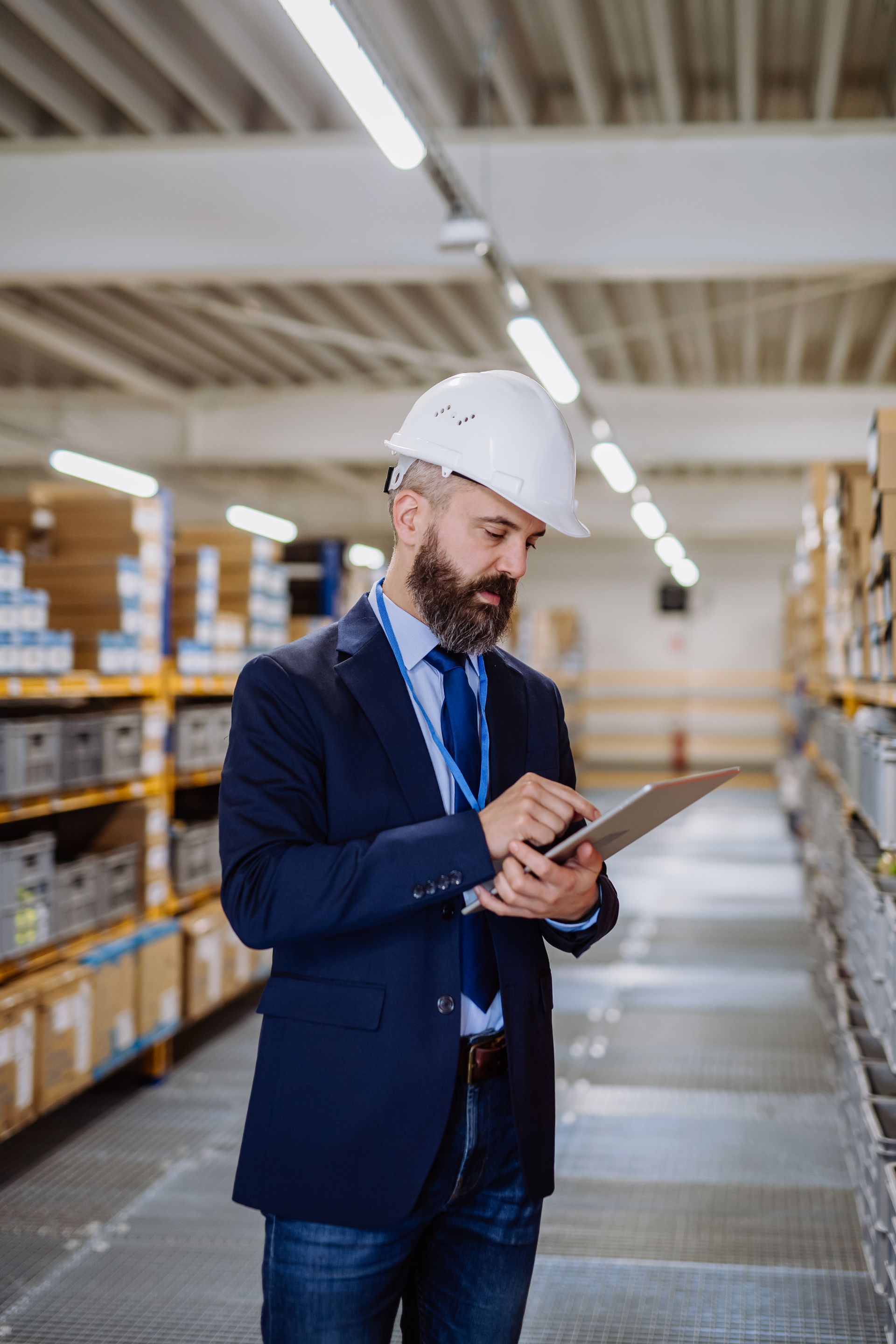 Man in a hard hat and suit using a tablet in a warehouse aisle with shelves of boxes
