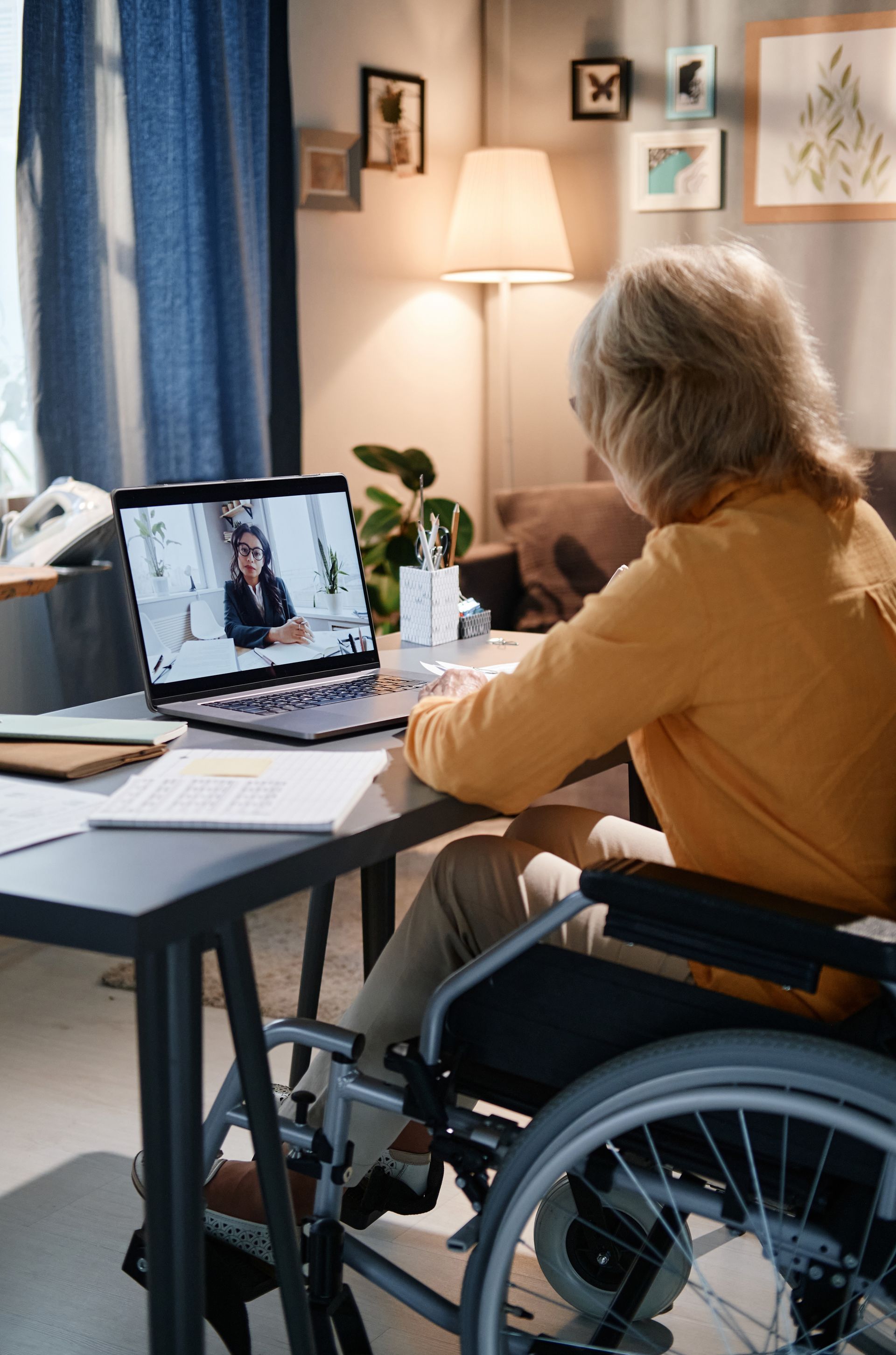 Person in a wheelchair video chatting at a home desk, facing a laptop in a cozy living room.