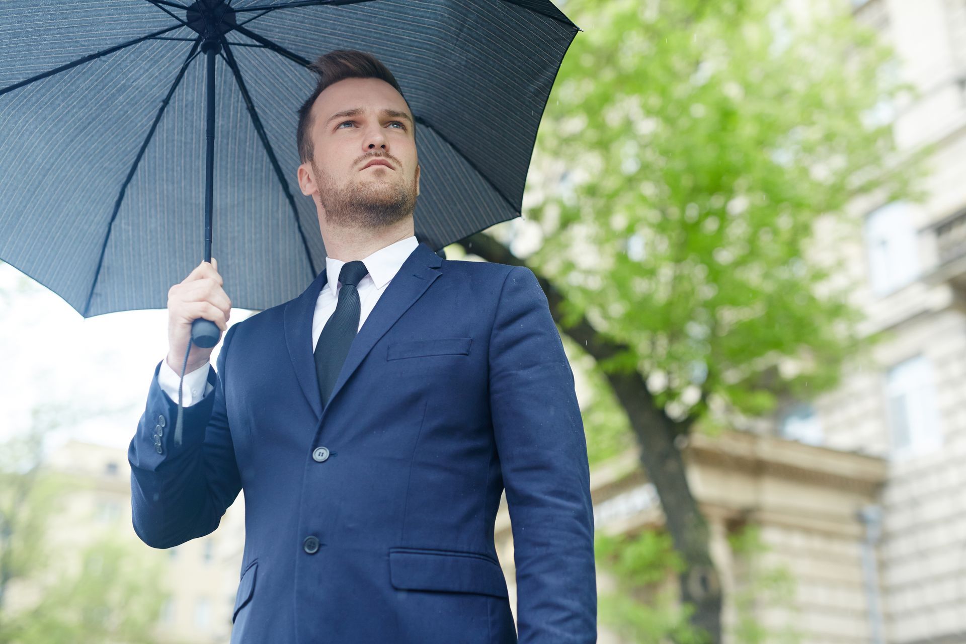 Man in a blue suit holding a black umbrella outdoors on a rainy day
