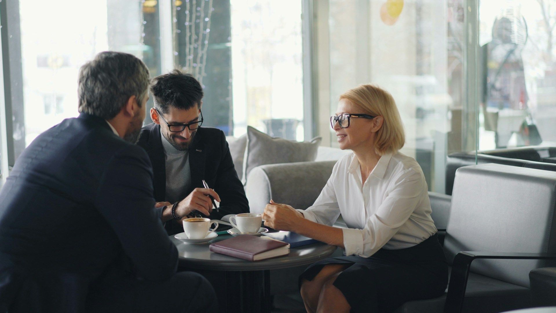 Three people in a bright office meeting, discussing over coffee at a table.