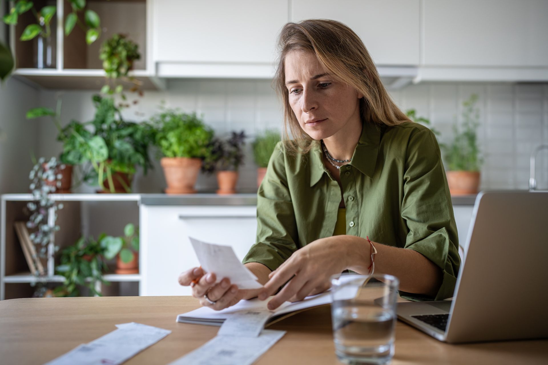 Woman reviewing papers at a desk with a laptop and glass of water in a bright home office