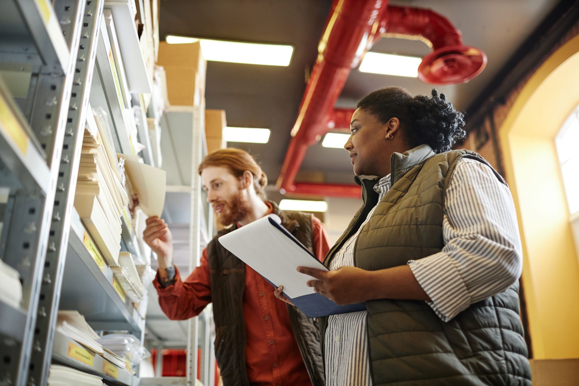 Two workers reviewing documents on warehouse shelves, one pointing upward and the other holding a clipboard.