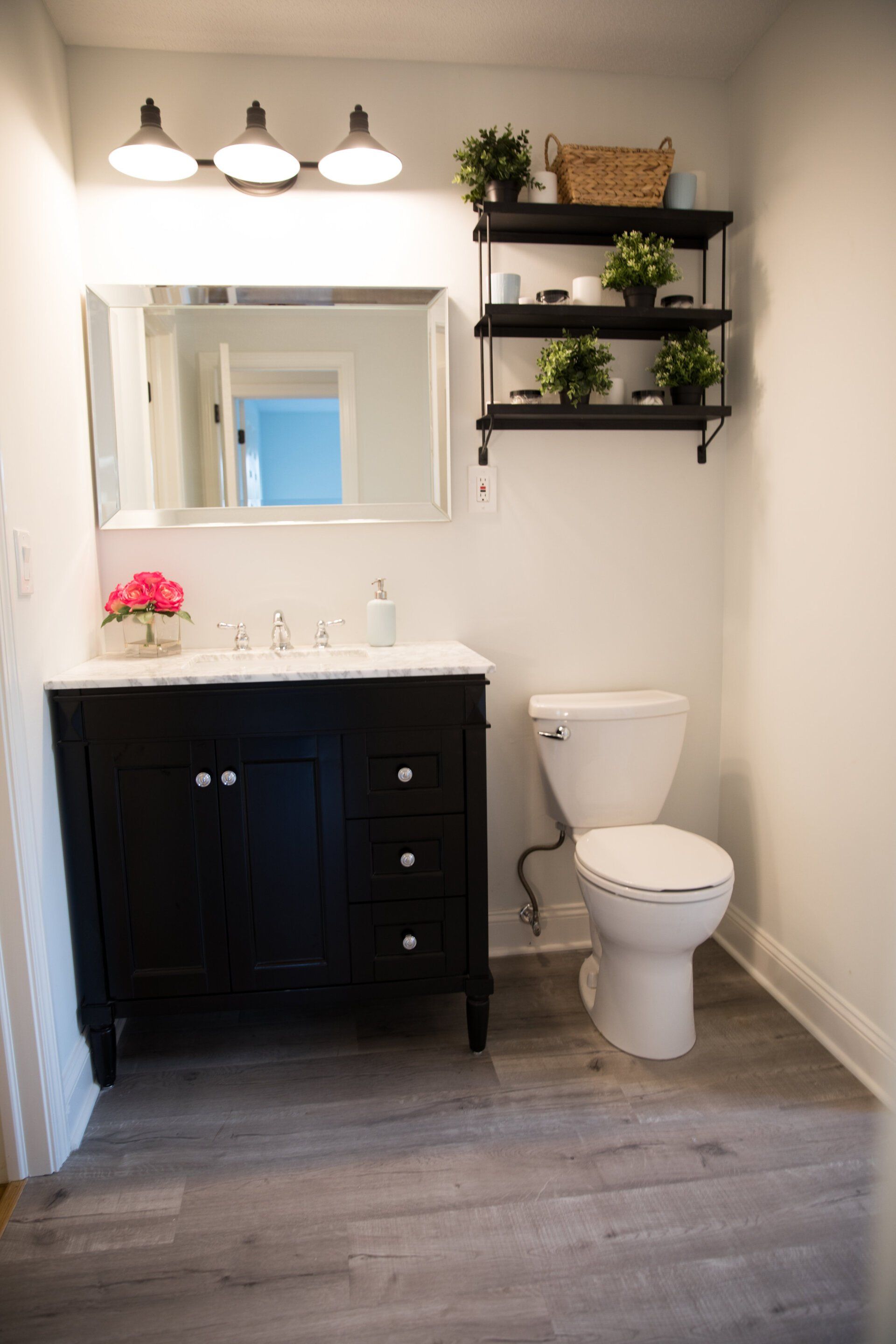 Bathroom with a black vanity, toilet, and shelves with plants, white walls, and gray floor.