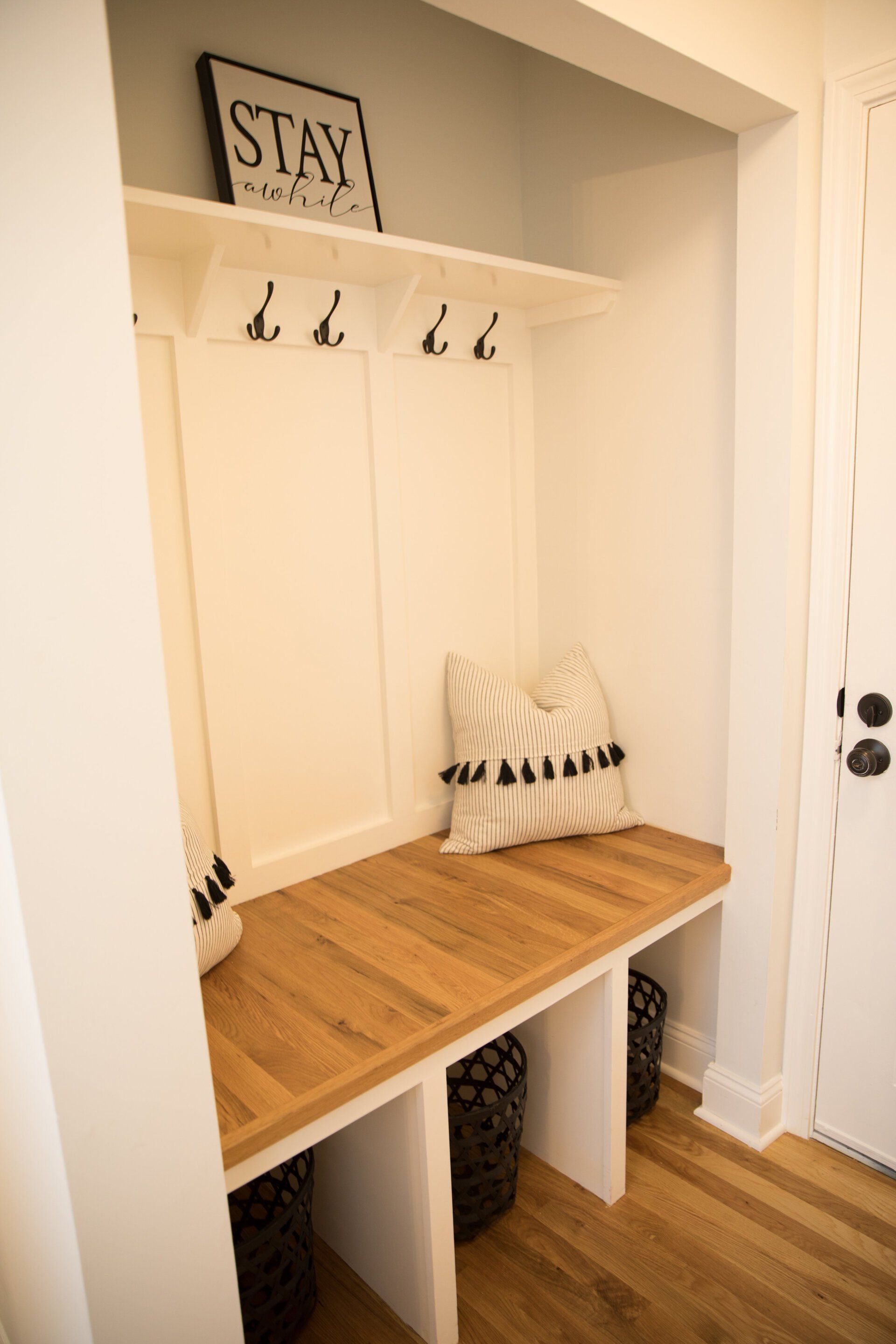 Built-in white mudroom bench with wooden seat, storage baskets, hooks, and decorative pillows.