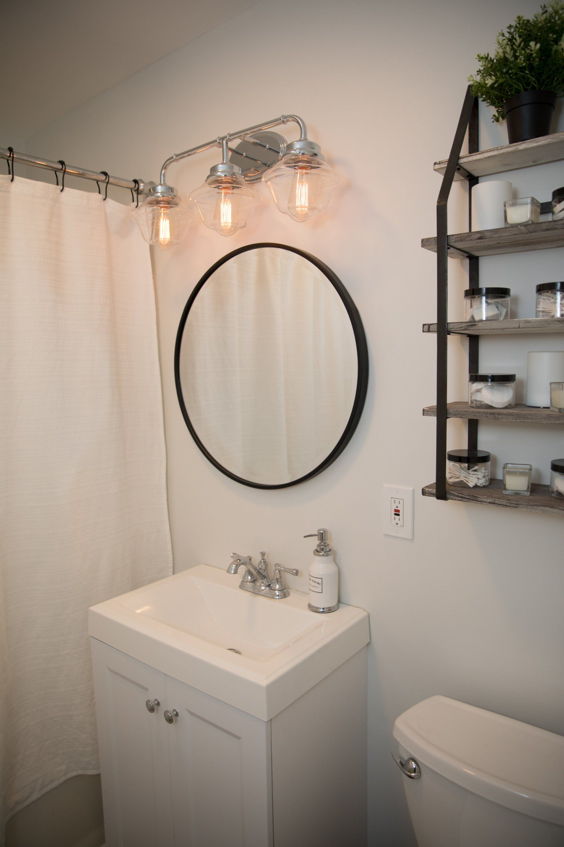 Bathroom with white vanity, round mirror, chrome light fixture, and open shelving unit.