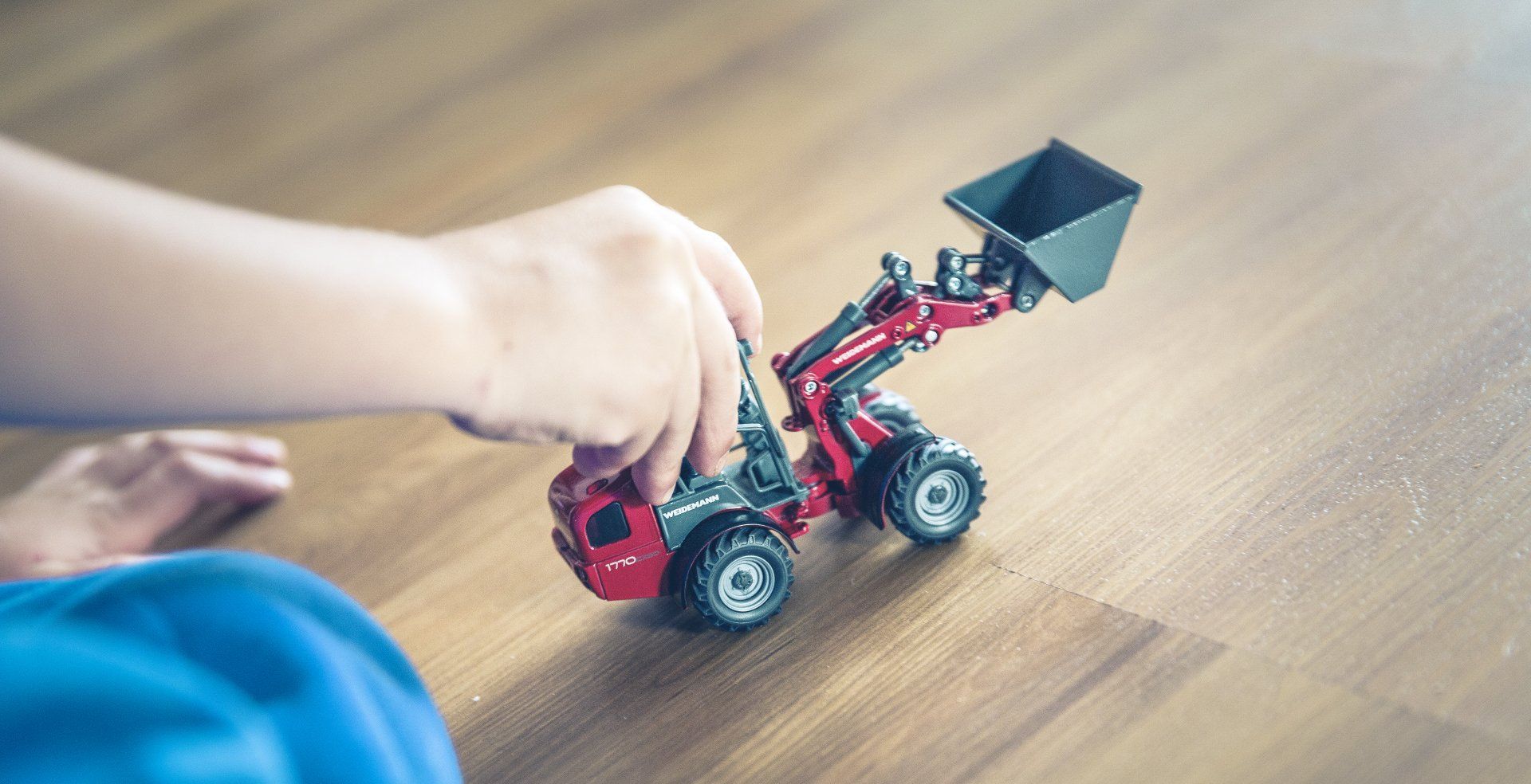 Child's hand playing with a red toy front-end loader on a wood floor.