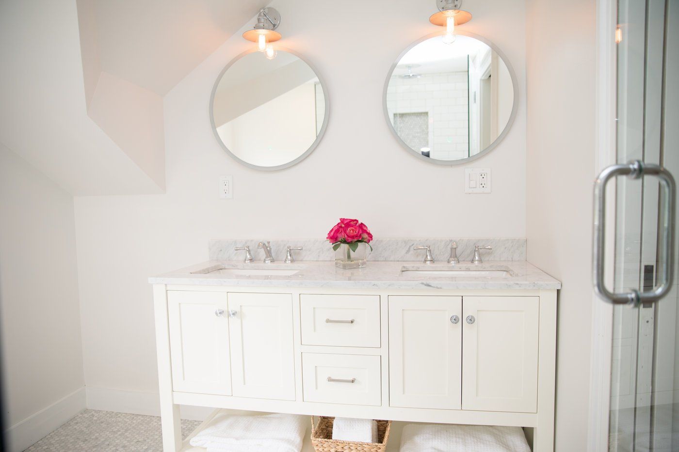 White bathroom with dual sinks, round mirrors, and a pink flower arrangement.
