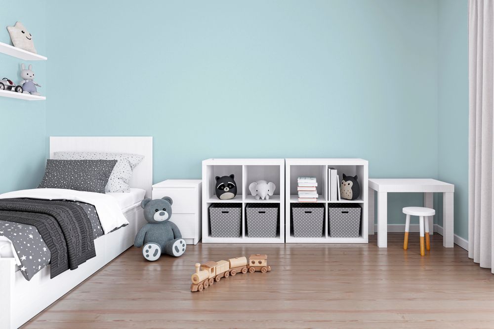Cameretta minimalista per bambini con letto bianco, contenitori per giocattoli e orsacchiotto su una parete azzurra.