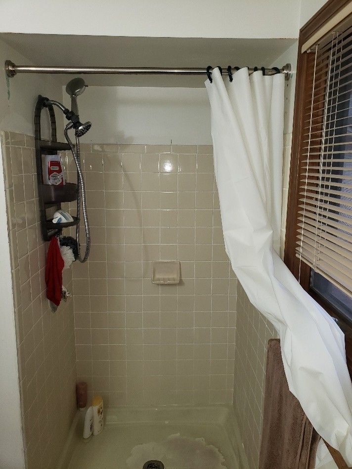 Shower with beige tiled walls, white curtain, and a window with blinds.