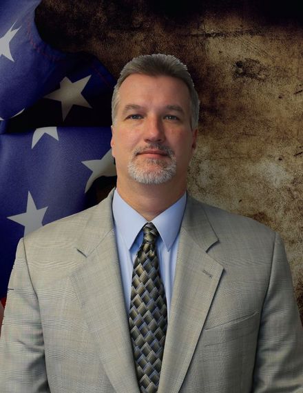 Man in a gray suit and tie posed before an American flag and textured brown backdrop.