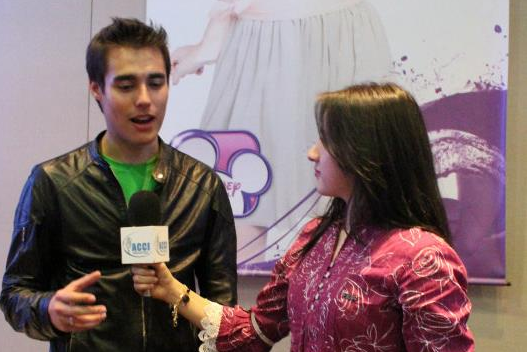 Un hombre con chaqueta negra es entrevistado por una mujer con un micrófono. Fondo morado.