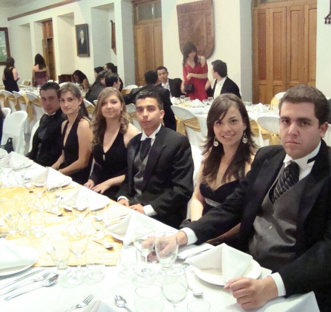 Personas vestidas de gala en una mesa de banquete. Algunos sonríen, otros miran a la cámara. Decoración dorada y blanca.