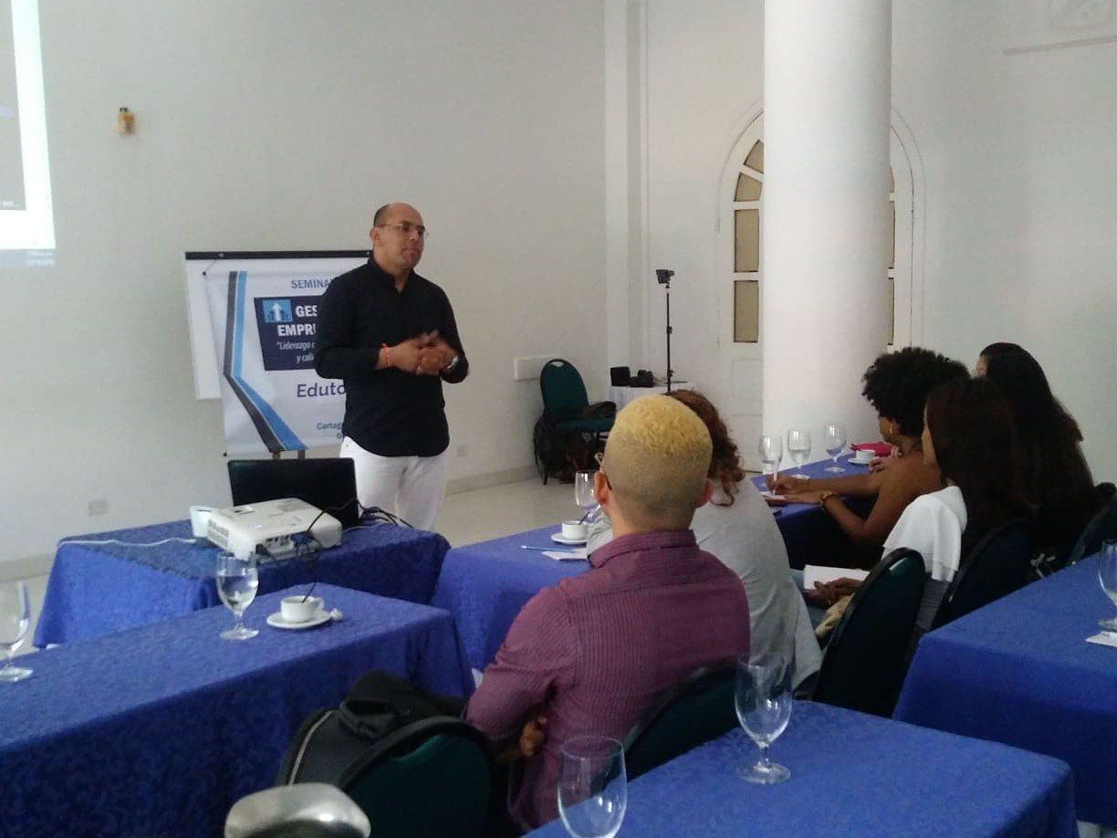 Hombre dando una presentación a una pequeña audiencia en una habitación luminosa con manteles azules.