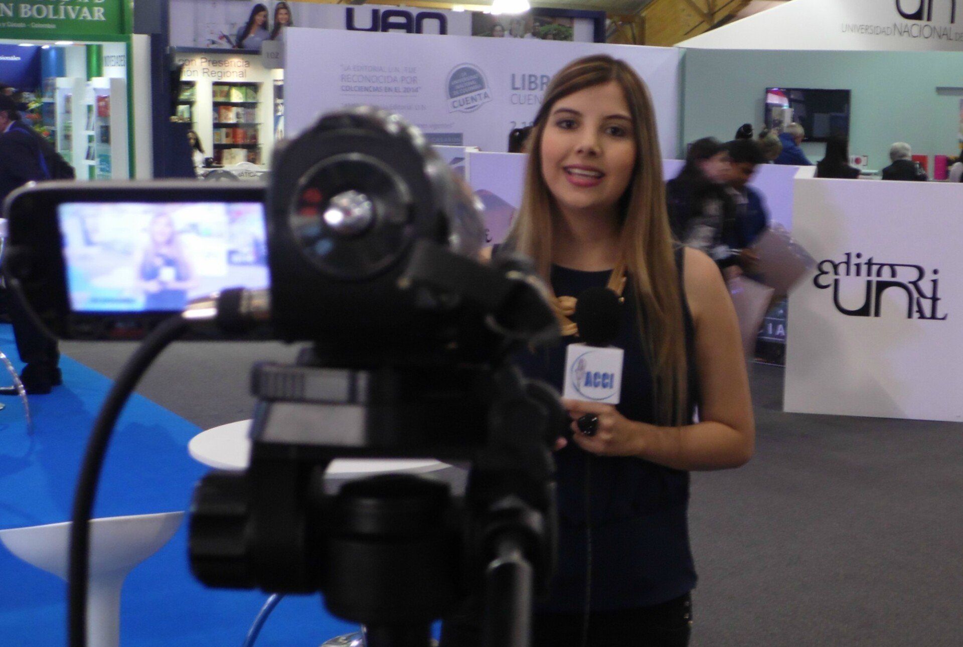 Mujer informando con una cámara de vídeo en una conferencia; está sonriendo y sosteniendo un micrófono.