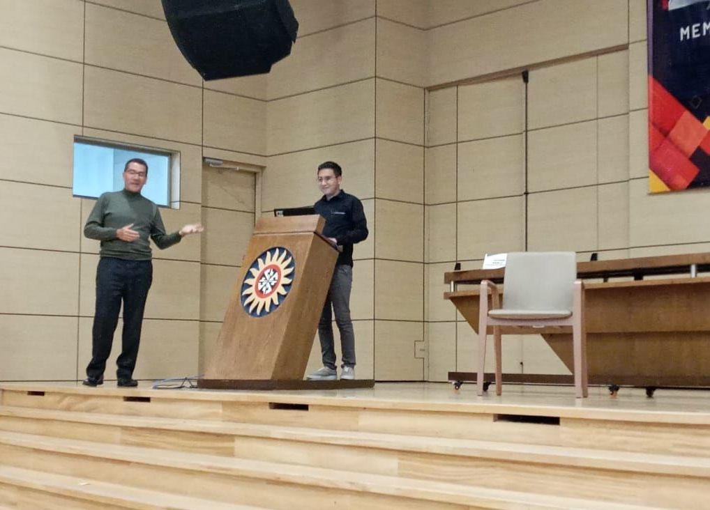 Dos hombres en un podio en un escenario dando una presentación; paneles de madera en el fondo.