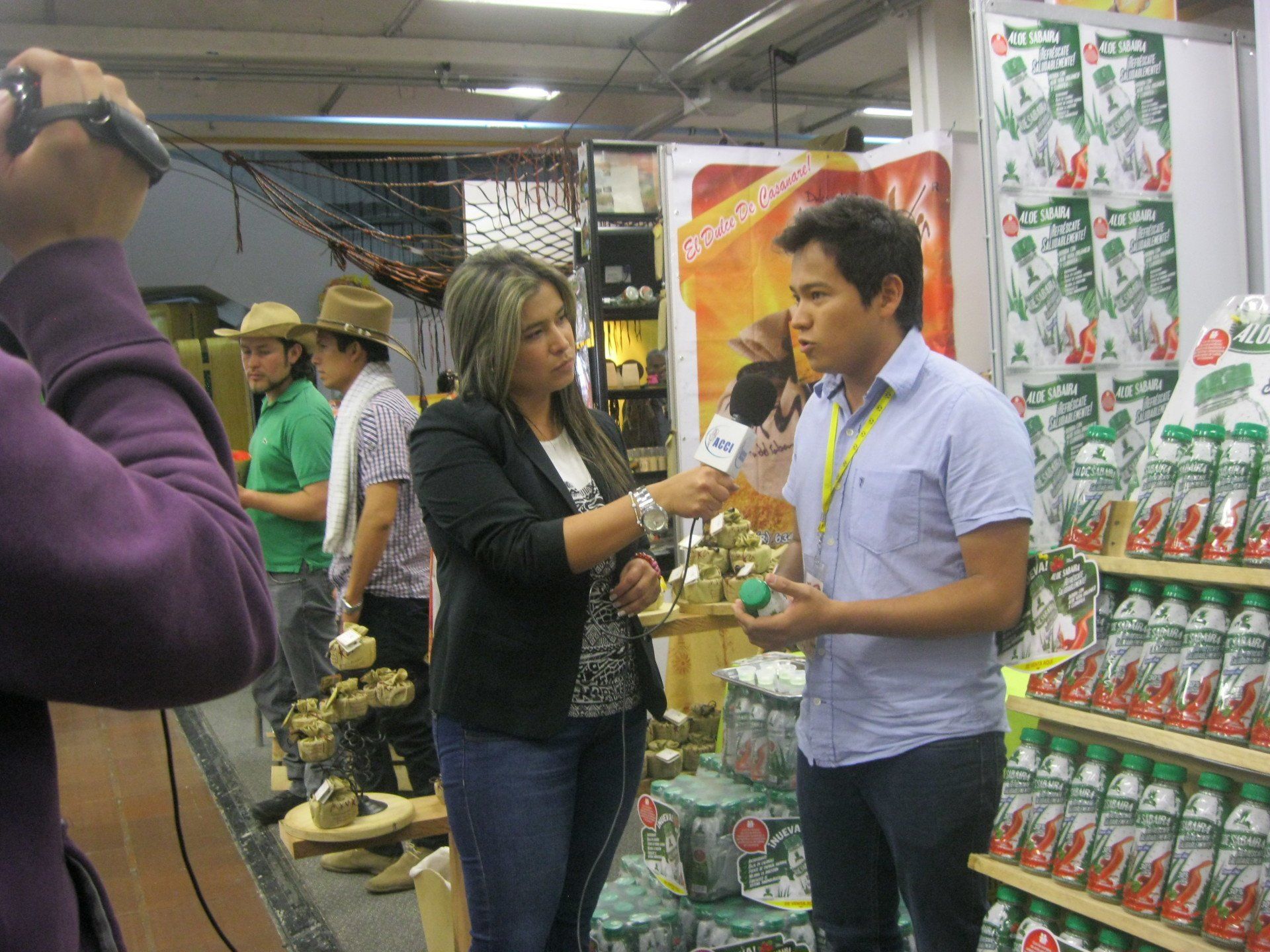 Un reportero entrevista a un hombre en un puesto con productos en estanterías. Al fondo, se ven otras personas y una cámara.