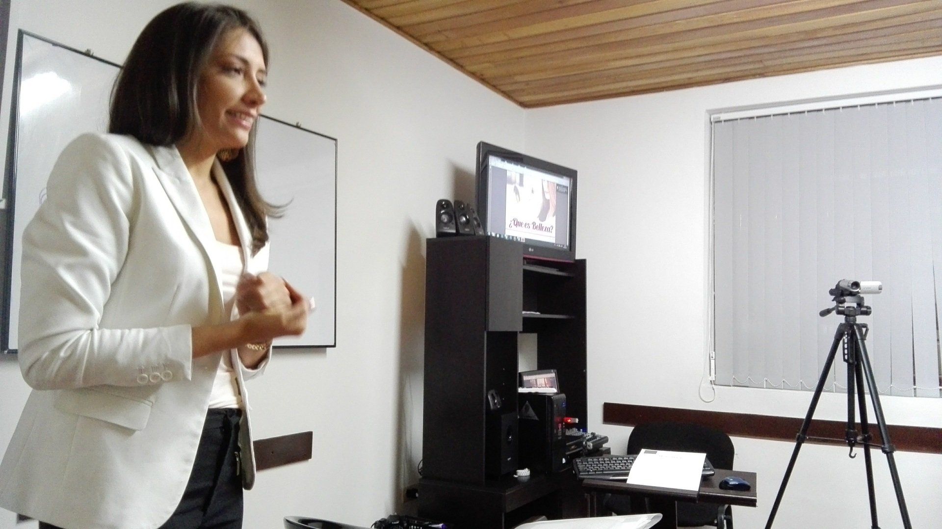 Una mujer con una chaqueta blanca habla en una habitación con una cámara en un trípode, una pizarra y un monitor.