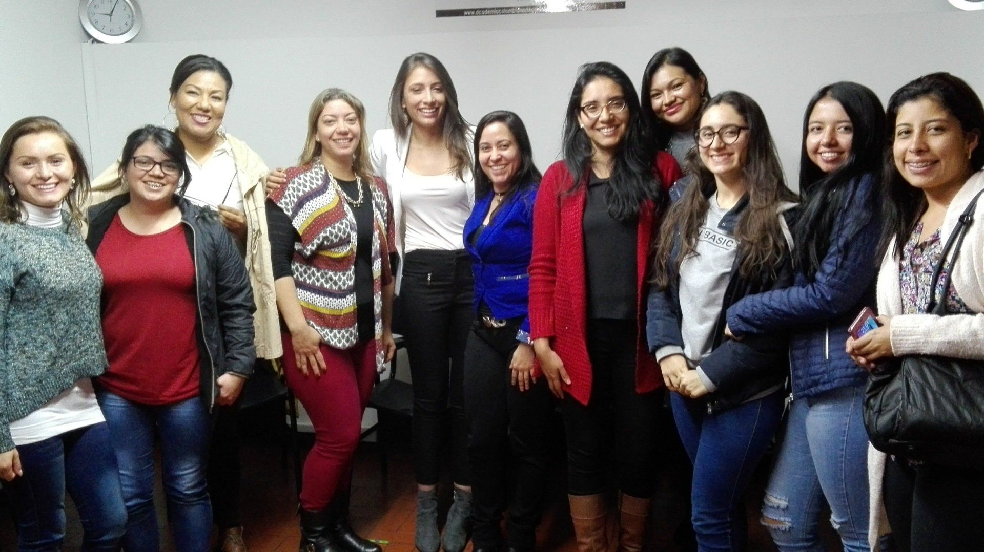 Grupo de mujeres sonriendo juntas en una habitación; algunas llevan chaquetas, tops y jeans.
