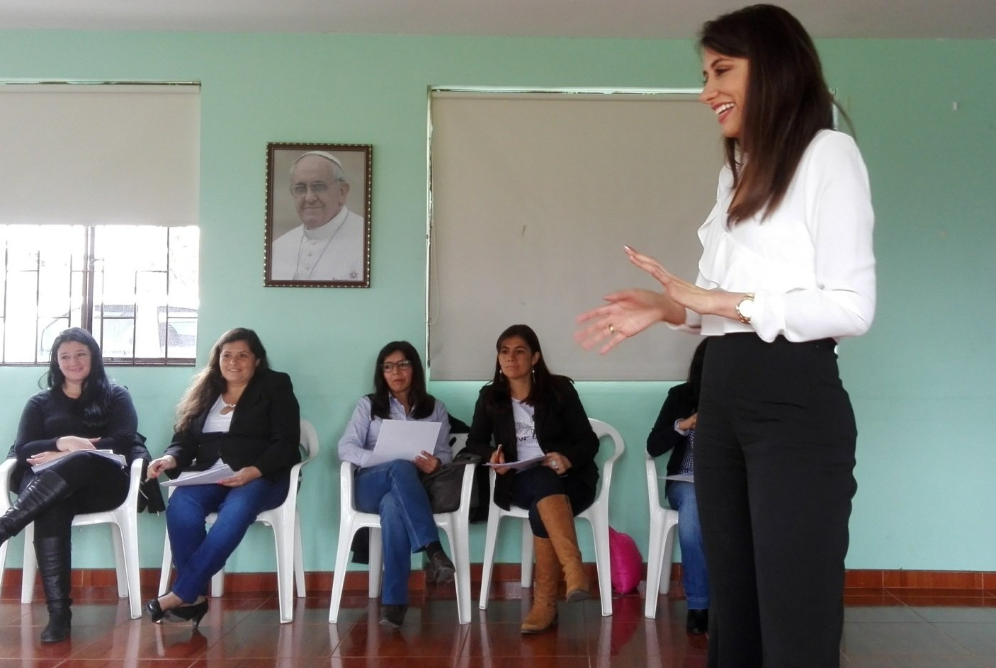 Mujer hablando a un grupo sentado en una habitación; un retrato es visible en la pared.