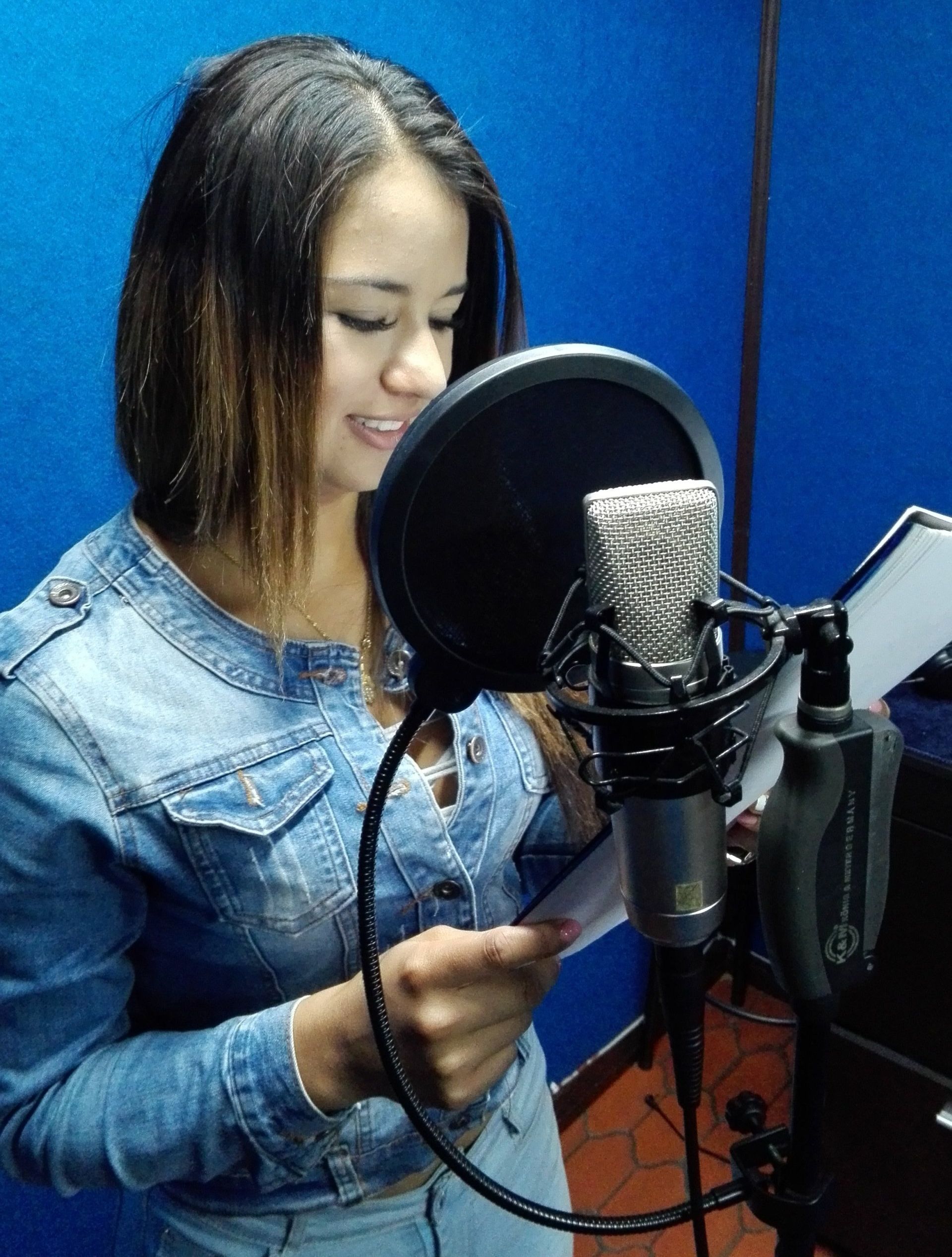 Mujer grabando voces en un estudio. Lleva una chaqueta vaquera y sonríe mientras sostiene un guion.