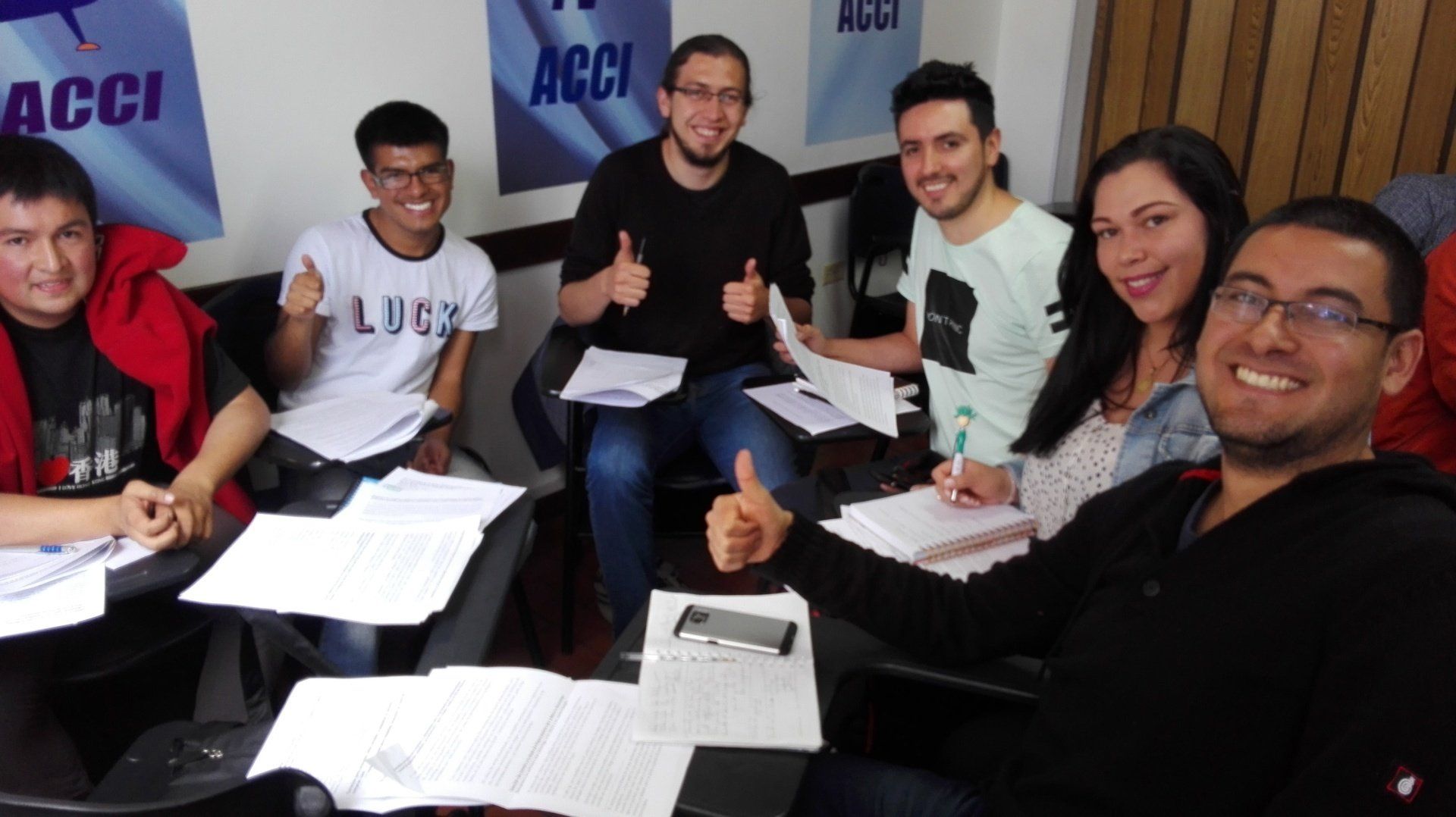 Grupo de personas en un aula, levantando el pulgar. Papeles y cuadernos sobre la mesa. El cartel de ACCI al fondo.