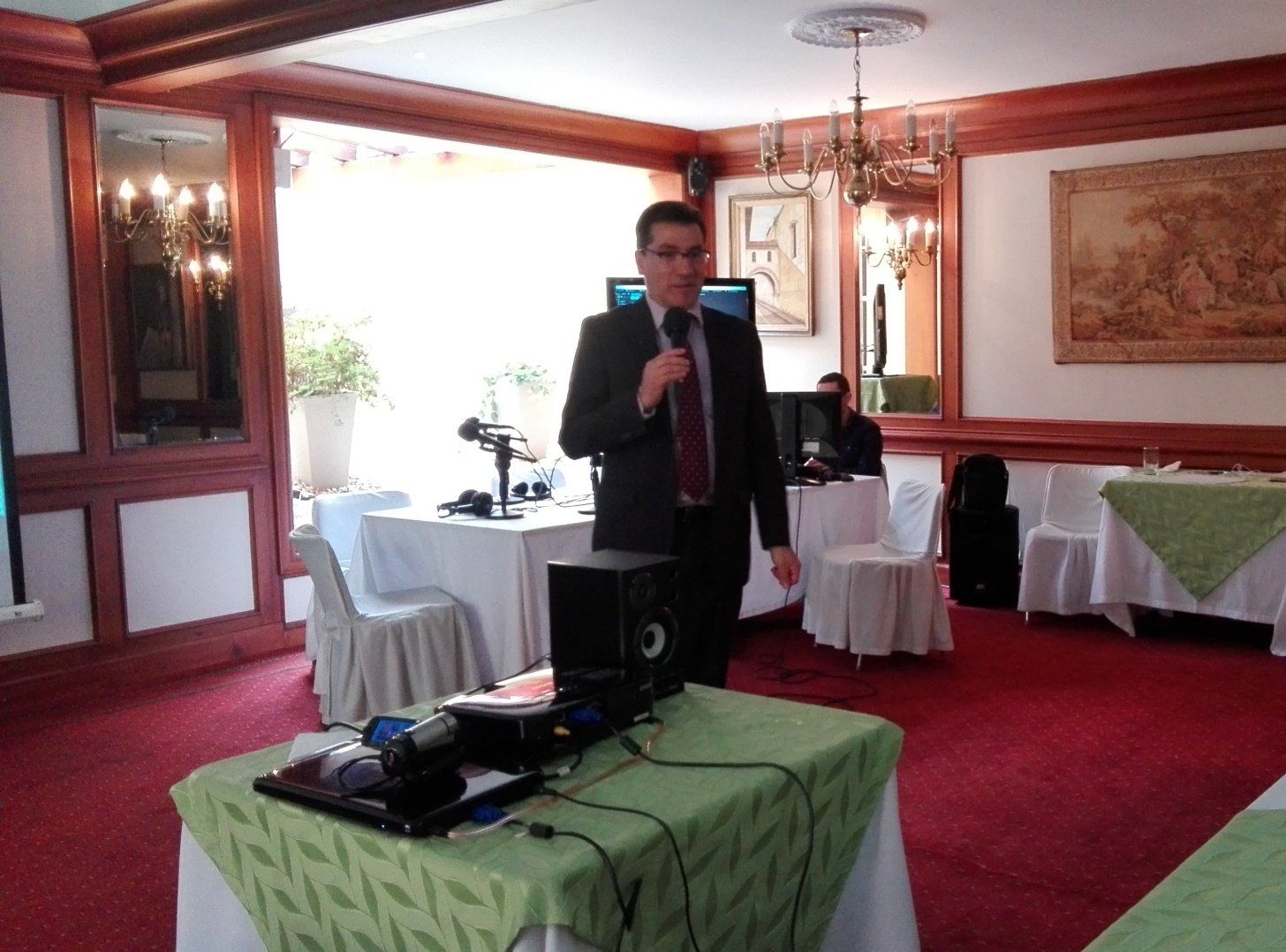 Un hombre de traje habla por un micrófono en un evento, con parlantes, una computadora portátil y mesas.