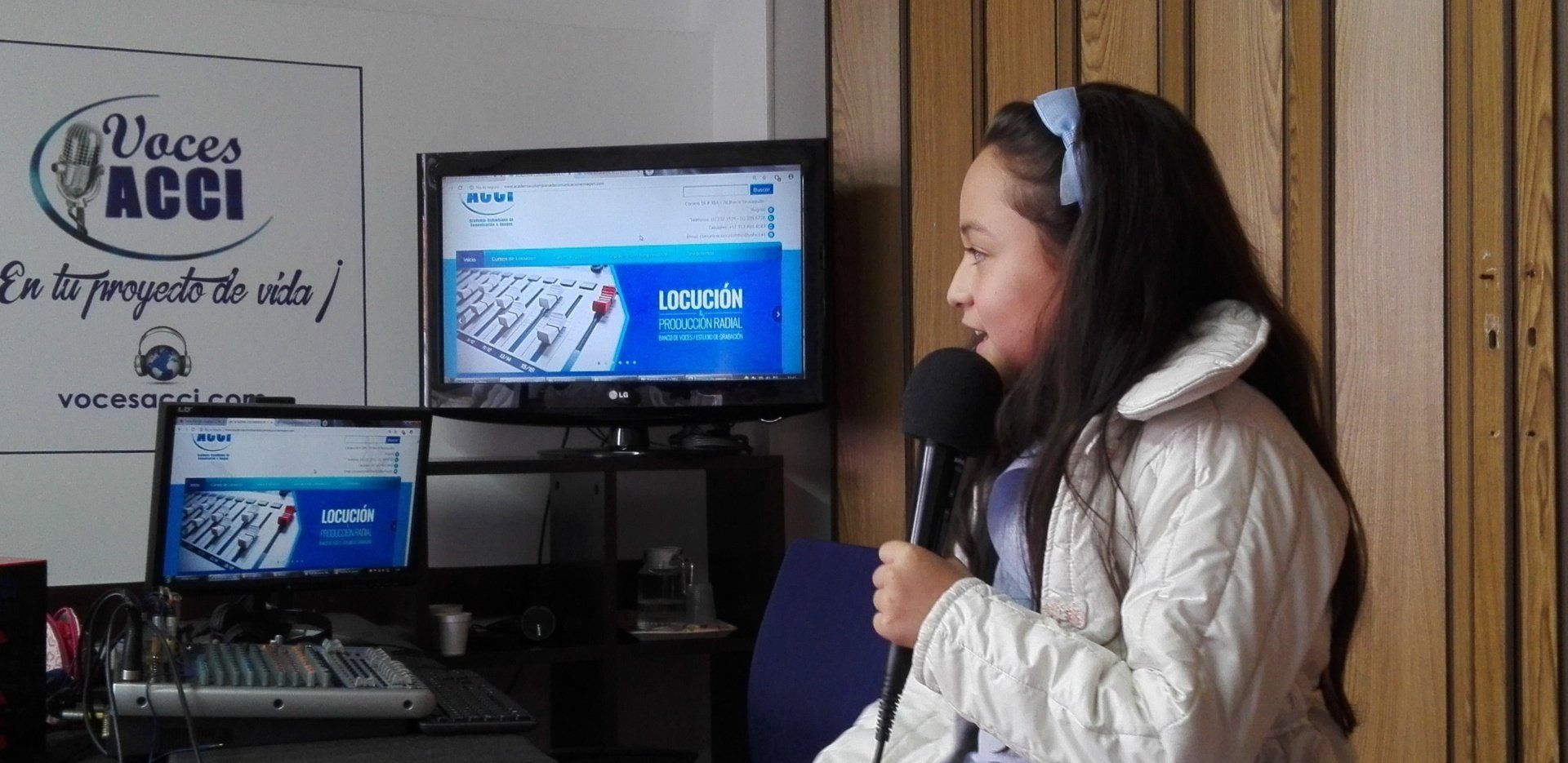 Chica hablando por un micrófono en un estudio con computadoras y un logo en la pared.