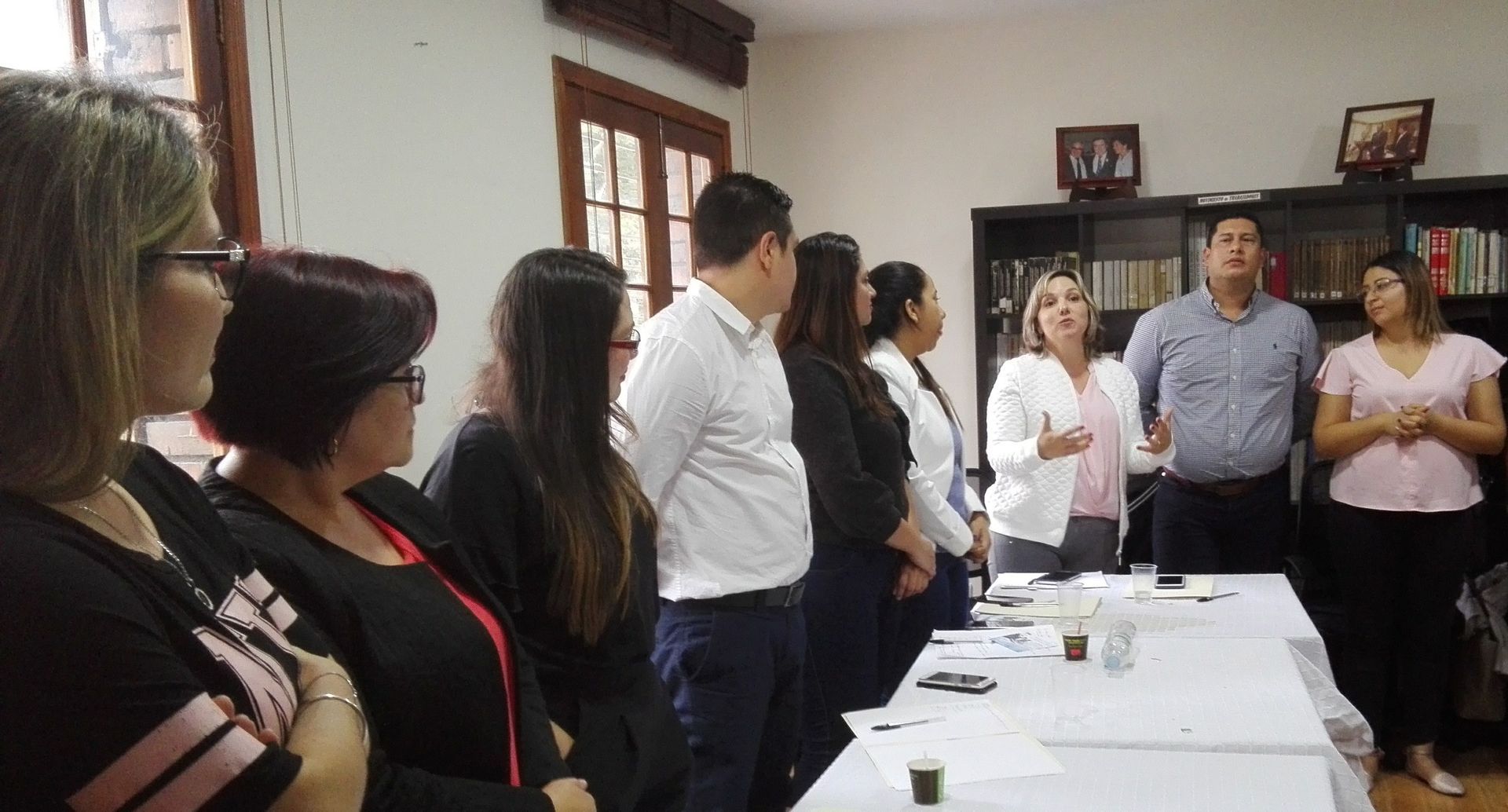 Grupo de personas en una oficina. Algunas están cerca de una mesa, otras junto a una estantería. Conversan.