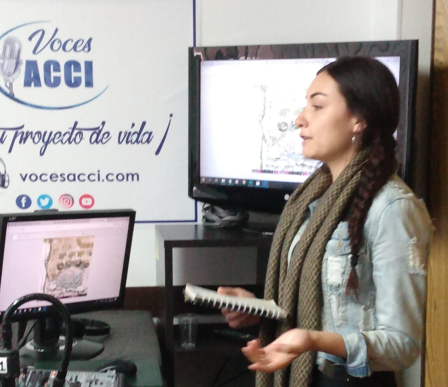 Mujer presentando en un estudio, con un cuaderno en la mano. Detrás de ella hay una pantalla y un logotipo.