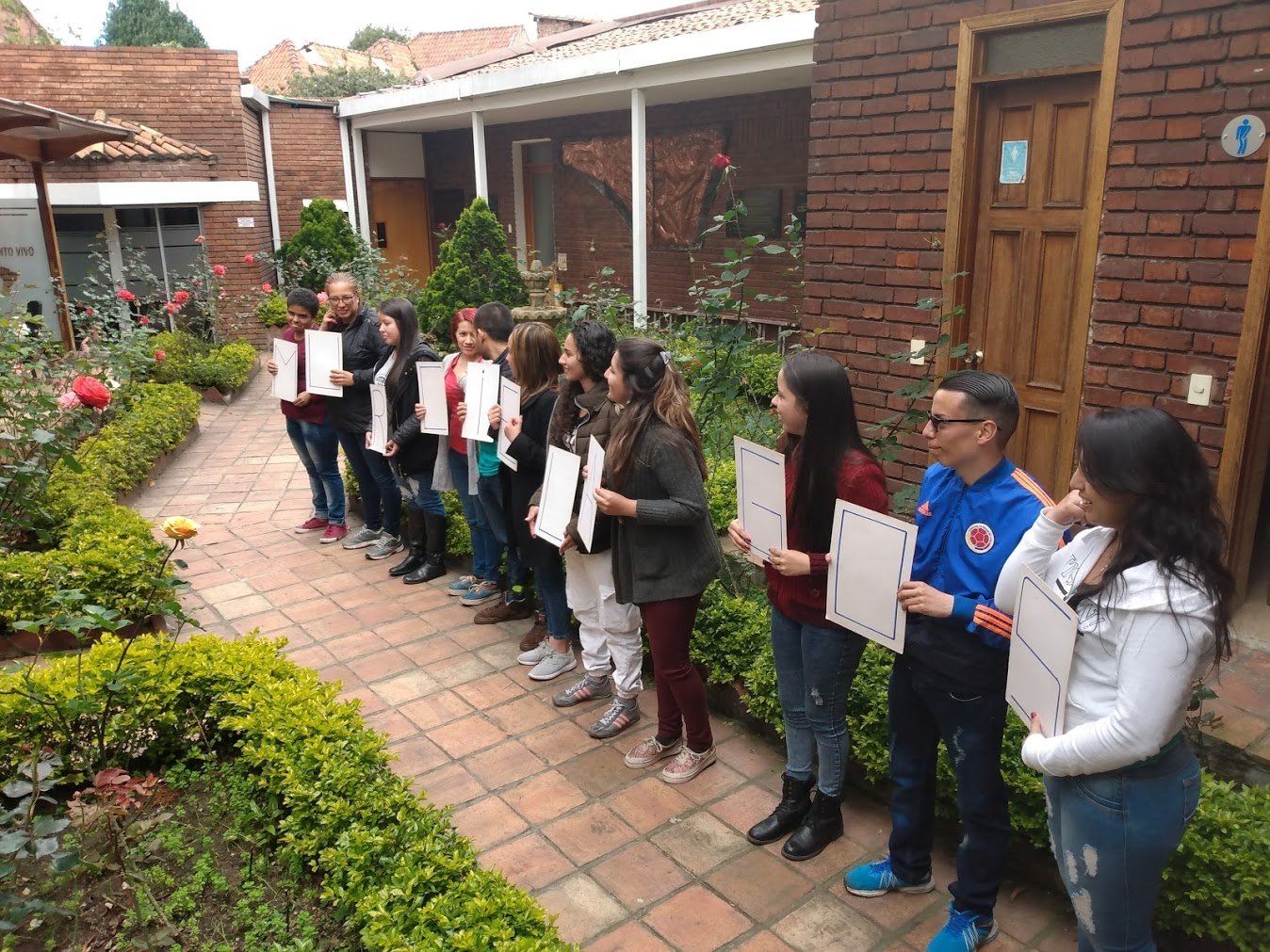 Grupo de personas sosteniendo papeles, de pie en un patio de ladrillo con vegetación y edificios.