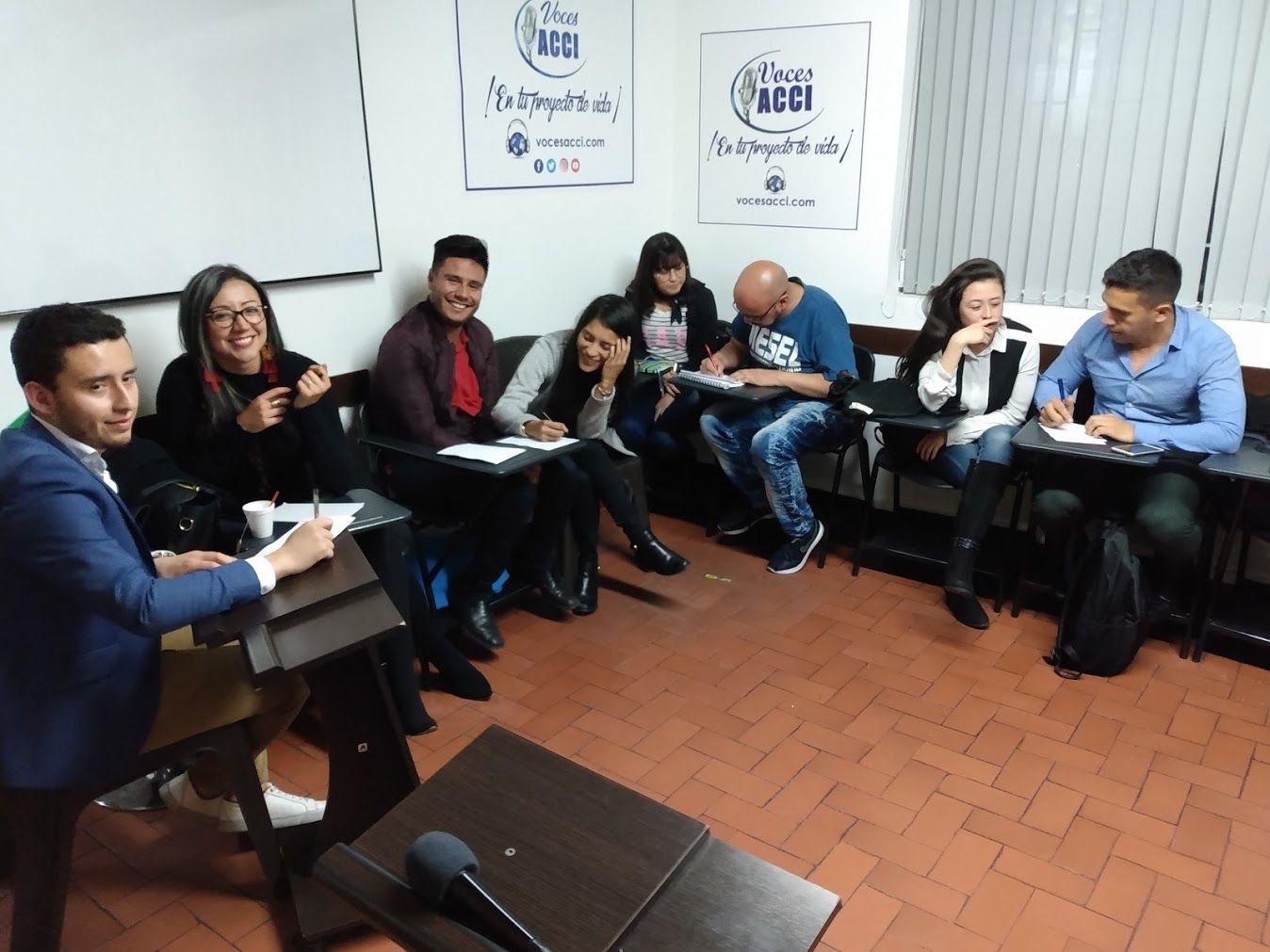 Personas en un aula, algunas escribiendo, otras escuchando. Vista interior con paredes blancas y suelo imitación madera.