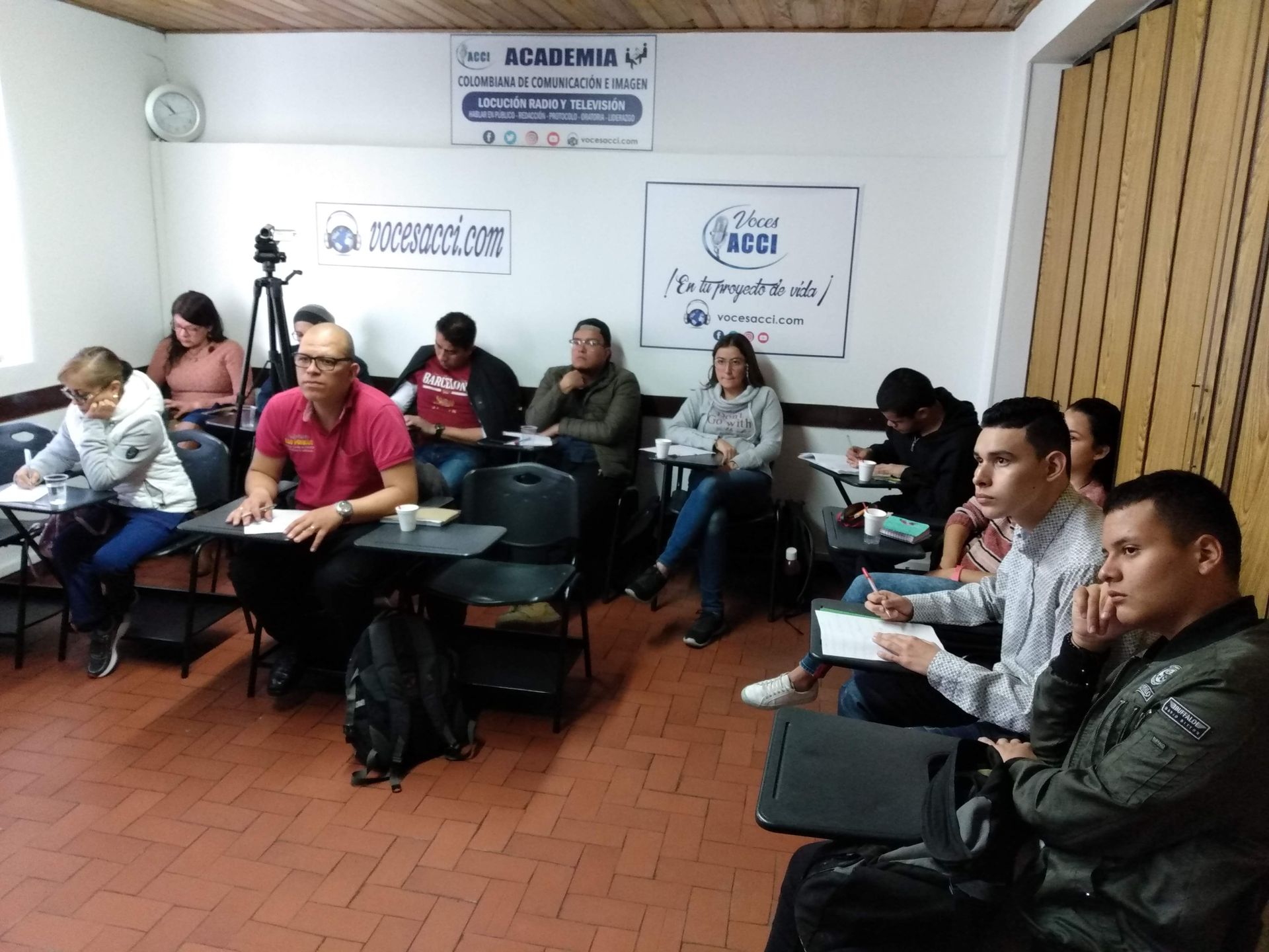 Un hombre presenta en una mesa a un grupo. Detrás hay una pared blanca. La gente toma notas.