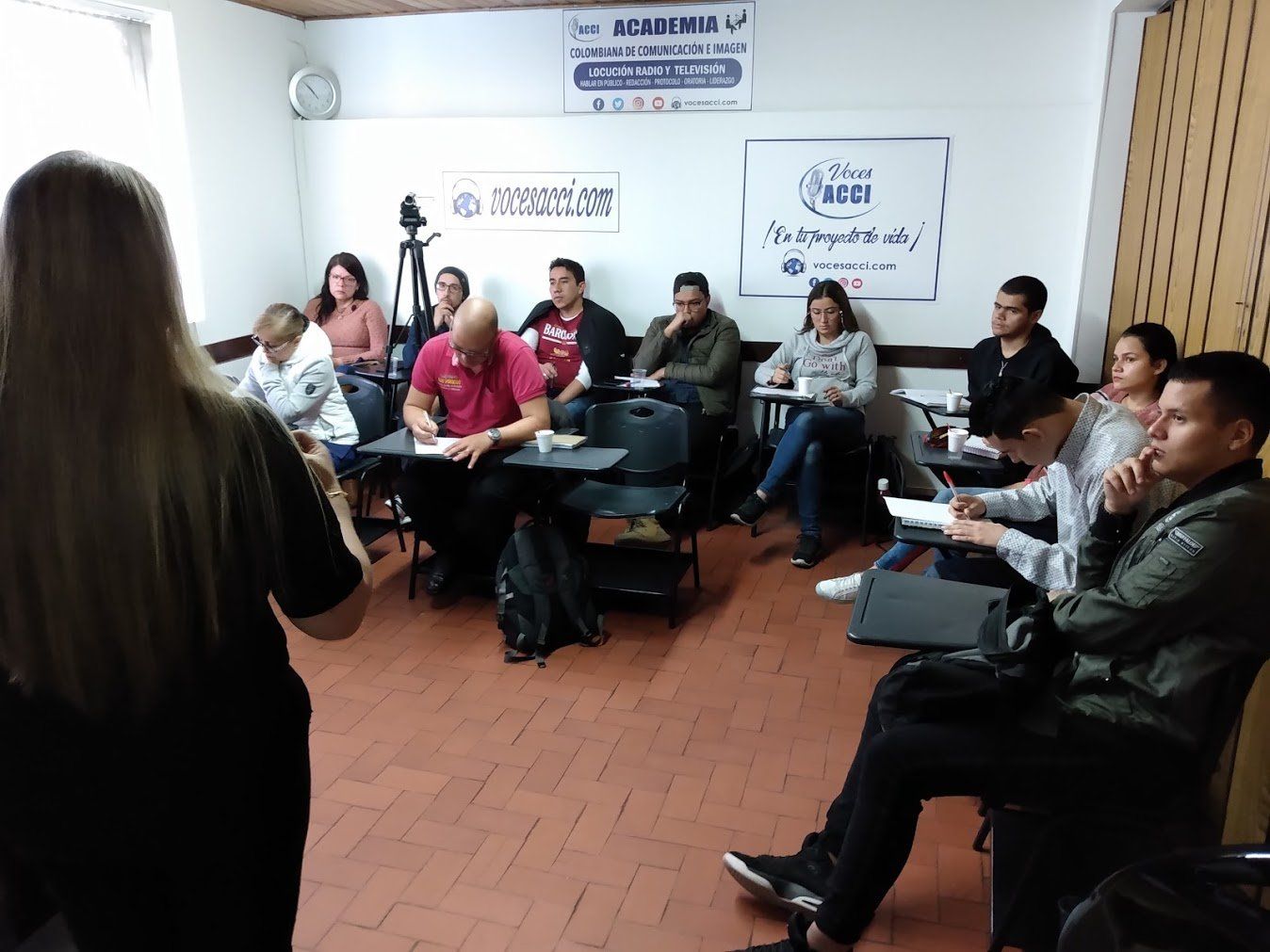 Una mujer da una conferencia a una clase de personas sentadas en escritorios en una sala con paredes blancas y logotipos.