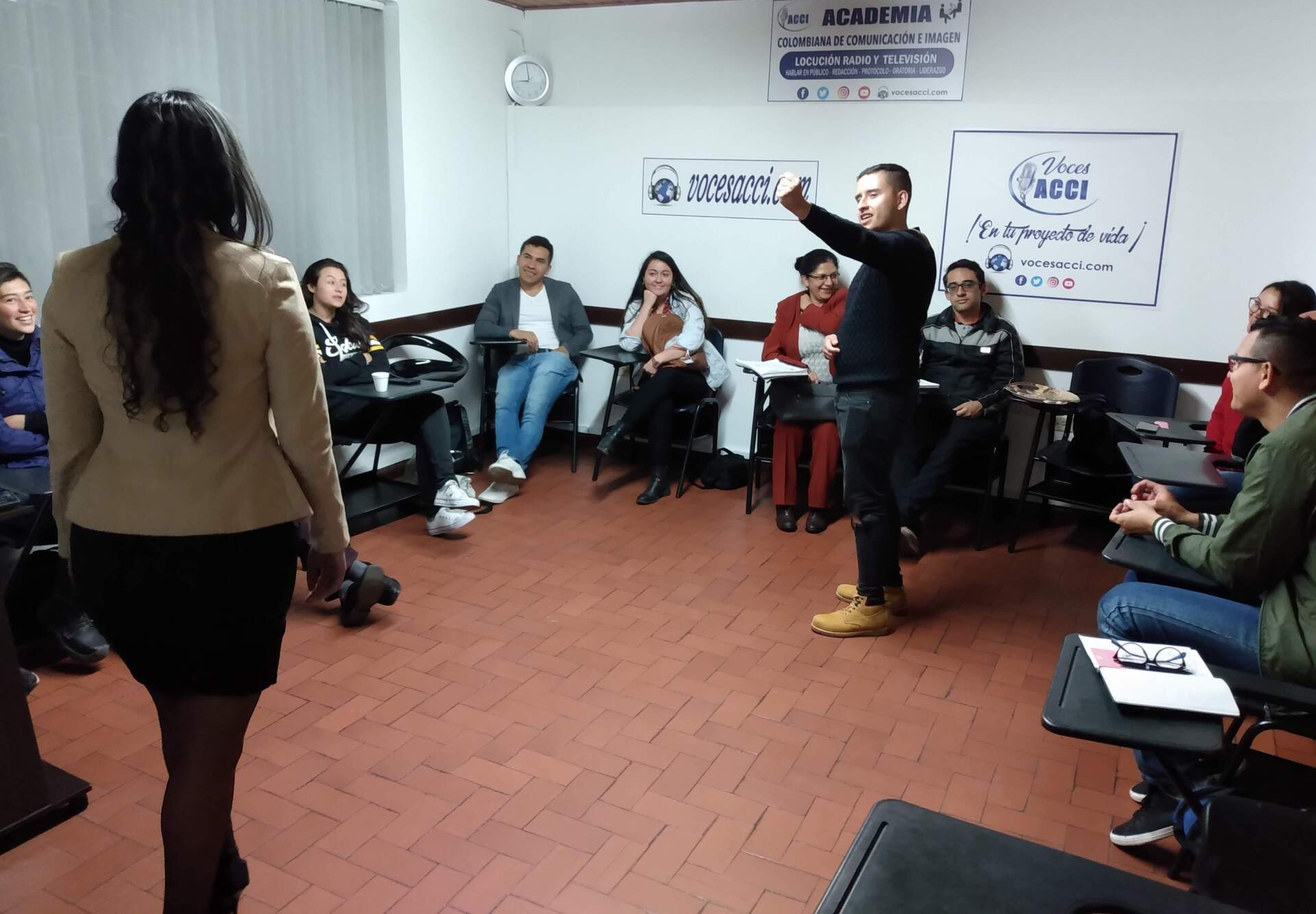 Un hombre gesticula en un aula. Los estudiantes están sentados, observando. Una mujer camina hacia ellos.