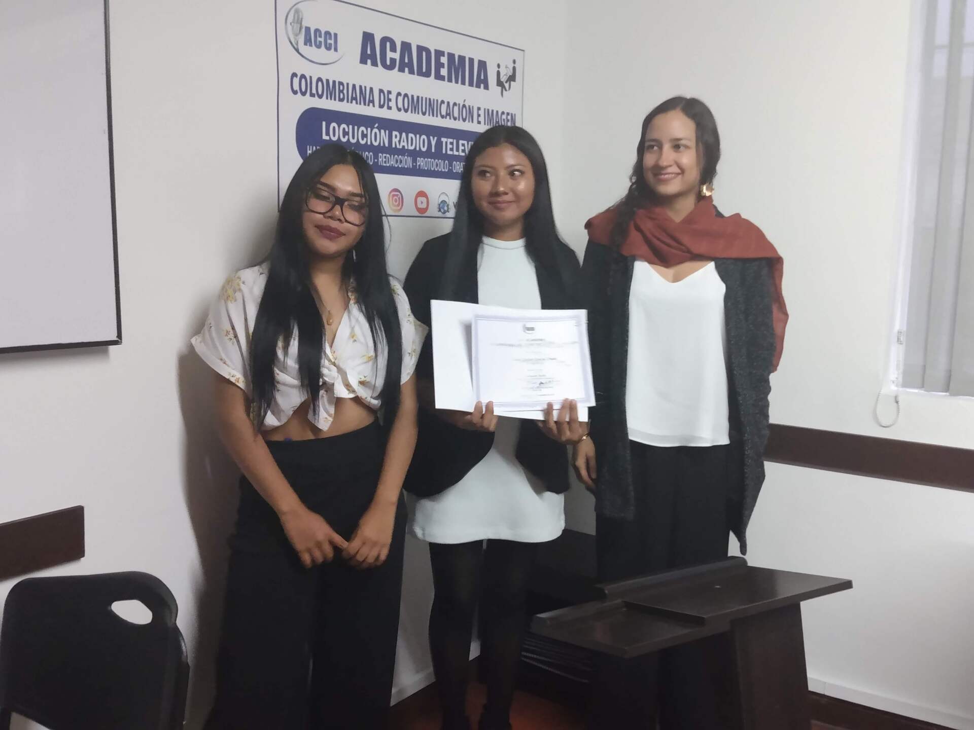 Tres mujeres en una oficina. Una sonríe con un certificado. Otras sonríen, de pie junto a un cartel.