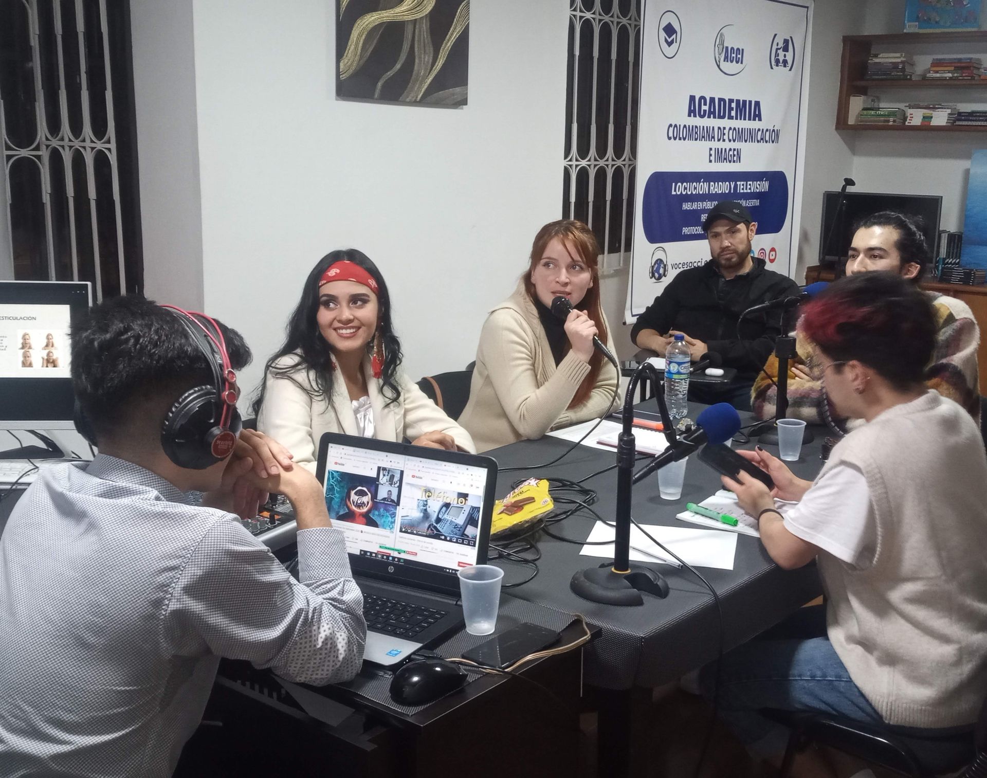 Personas grabando un podcast en una mesa con micrófonos, una computadora portátil que muestra una caricatura y auriculares.