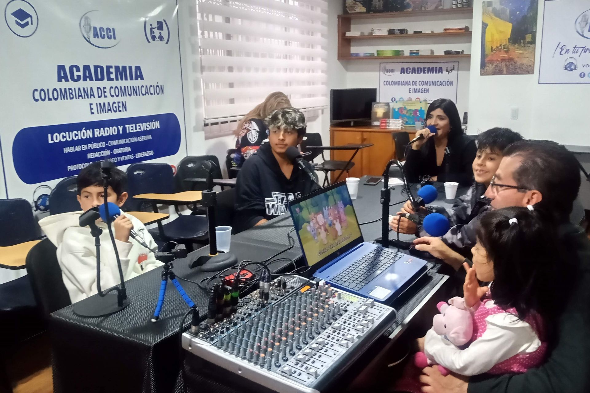 Personas en un estudio con micrófonos, una mesa de mezclas y una computadora portátil; pancarta en el fondo.