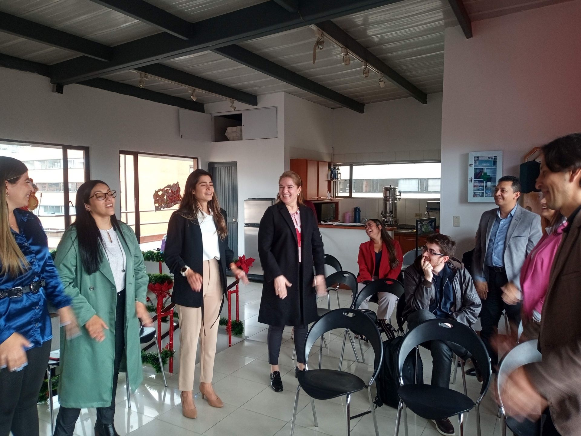 Personas en una sala con sillas, algunas de pie y gesticulando. Interior luminoso con una cocina visible al fondo.