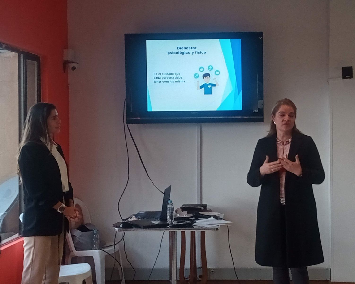 Dos mujeres se presentan en una sala con una pantalla que muestra una presentación. Una gesticula; la otra se queda cerca.