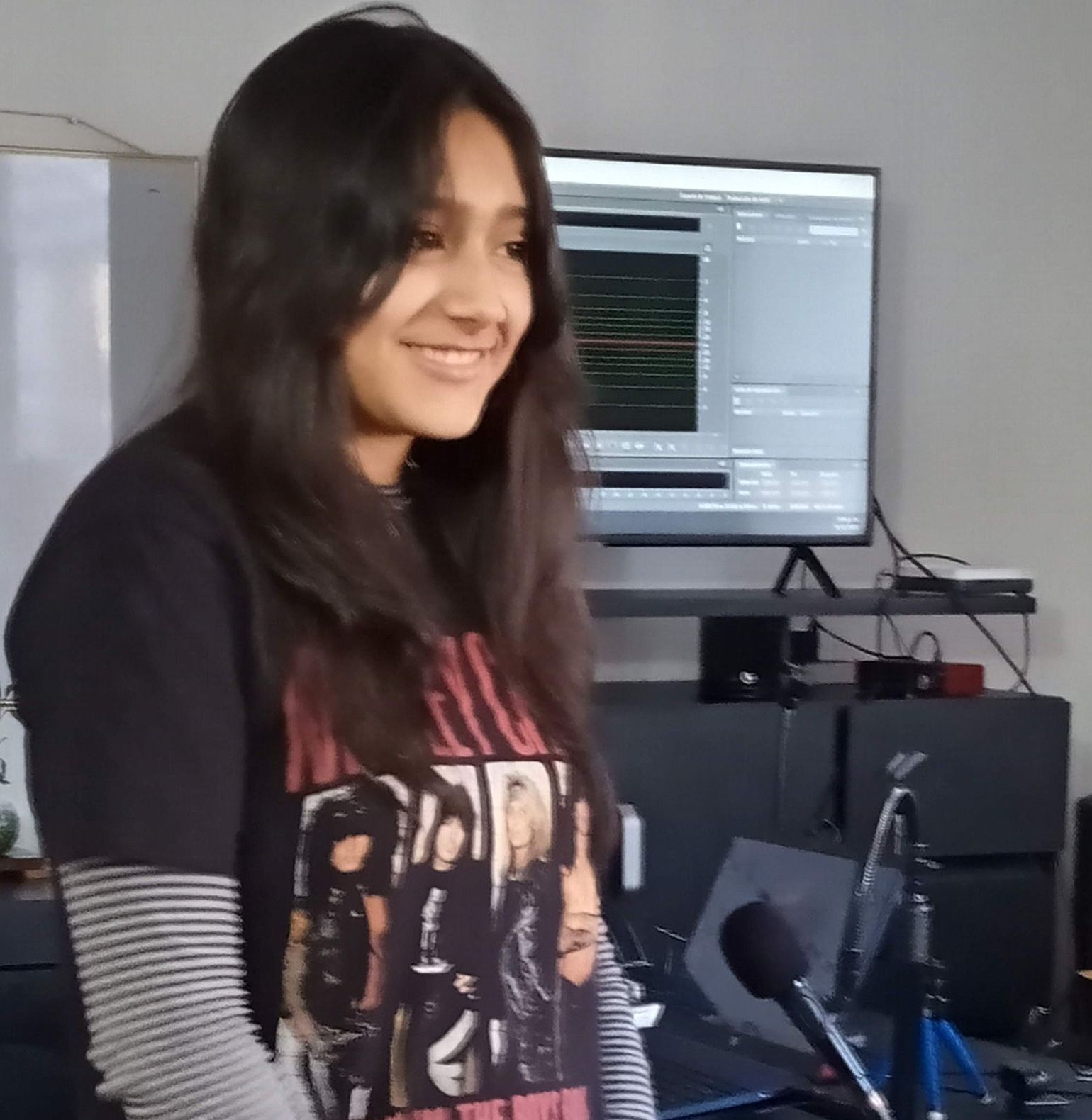 Mujer sonriendo, vistiendo una camiseta estampada y una camisa de manga larga a rayas, frente a una pantalla de computadora.