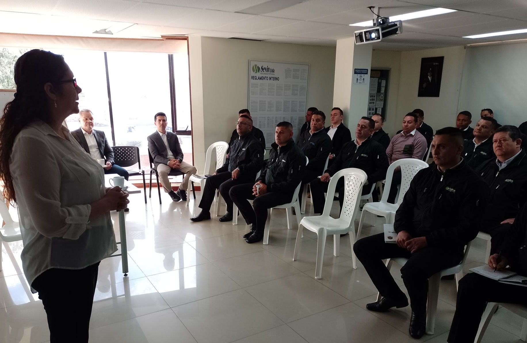 Una mujer habla con un grupo sentado, probablemente en una sala de conferencias. Personas uniformadas, posiblemente policías.