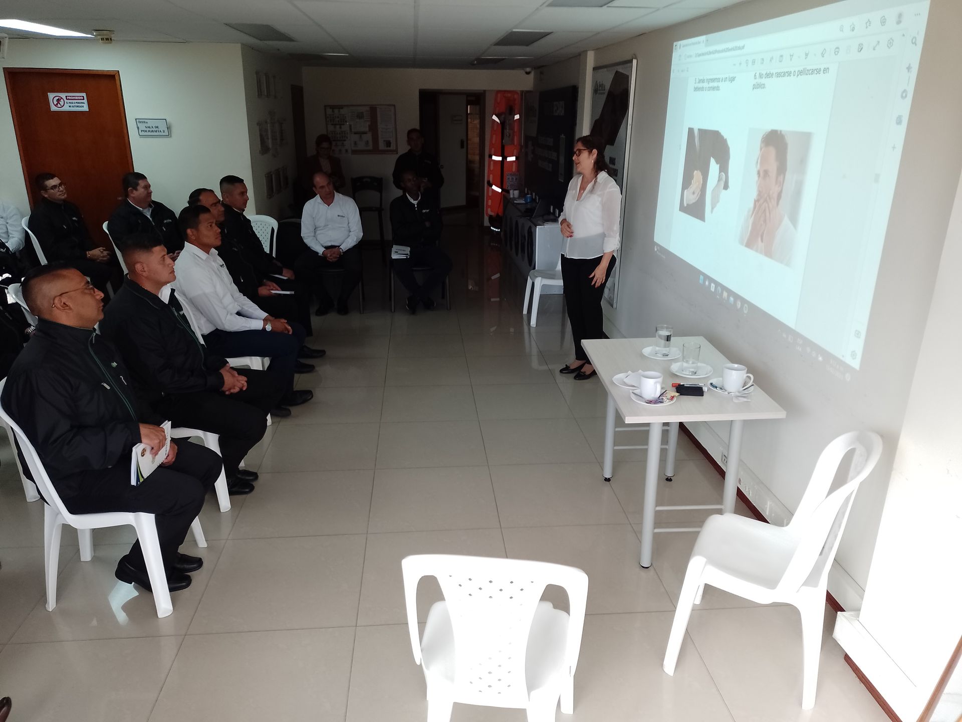 Una mujer presenta a un grupo sentado. Un proyector proyecta imágenes. Sillas y mesa blancas. Interior.