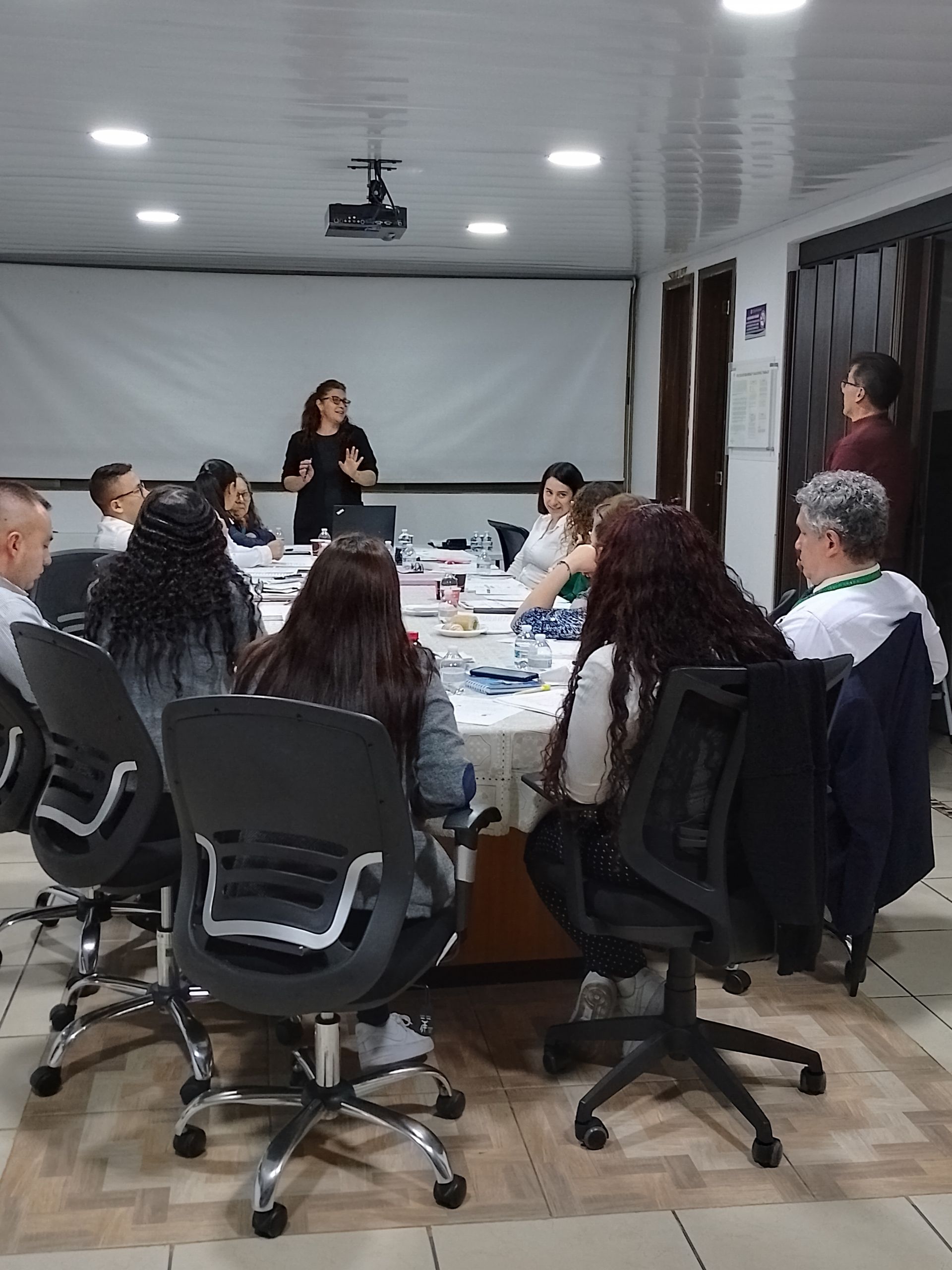 Personas en una sala de reuniones, una mujer presentando en la mesa, otros sentados escuchando.
