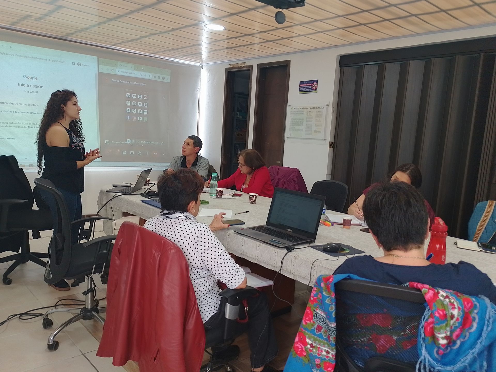 Mujer presentando en una mesa a varias personas, con computadora portátil y proyector en una sala de reuniones.